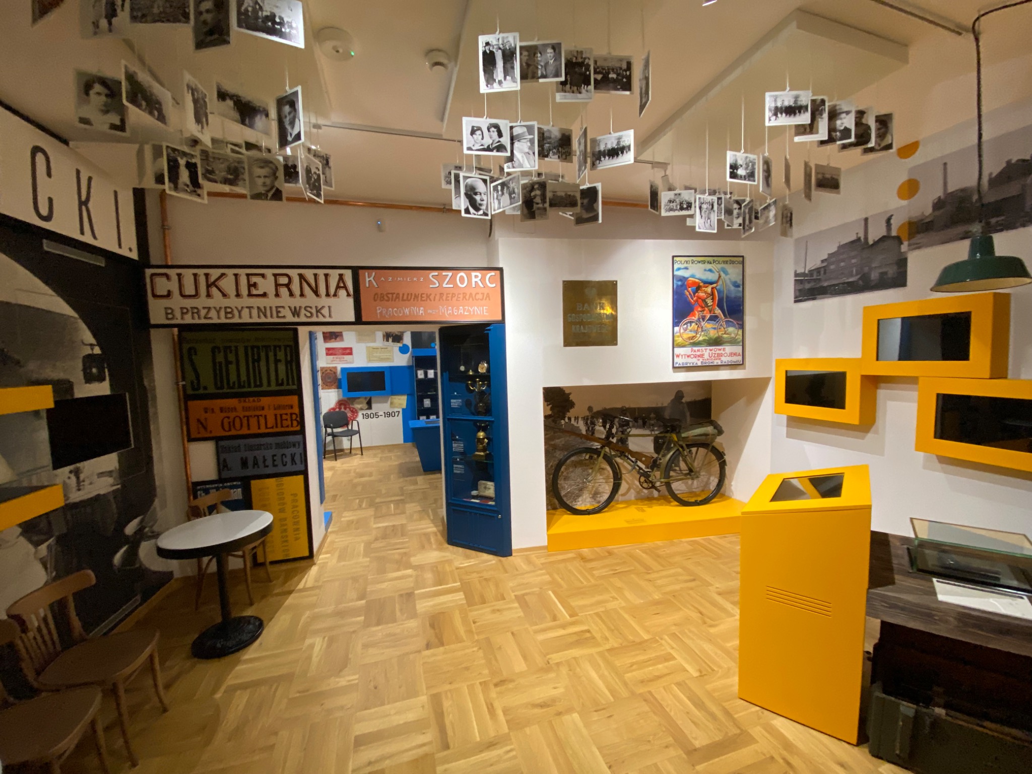 Wnętrze muzeum z ekspozycją historyczną: wiszące zdjęcia portretowe, repliki szyldów sklepowych i plakatów propagandowych na ścianach, eksponaty w gablotach oraz rower turystyczny.