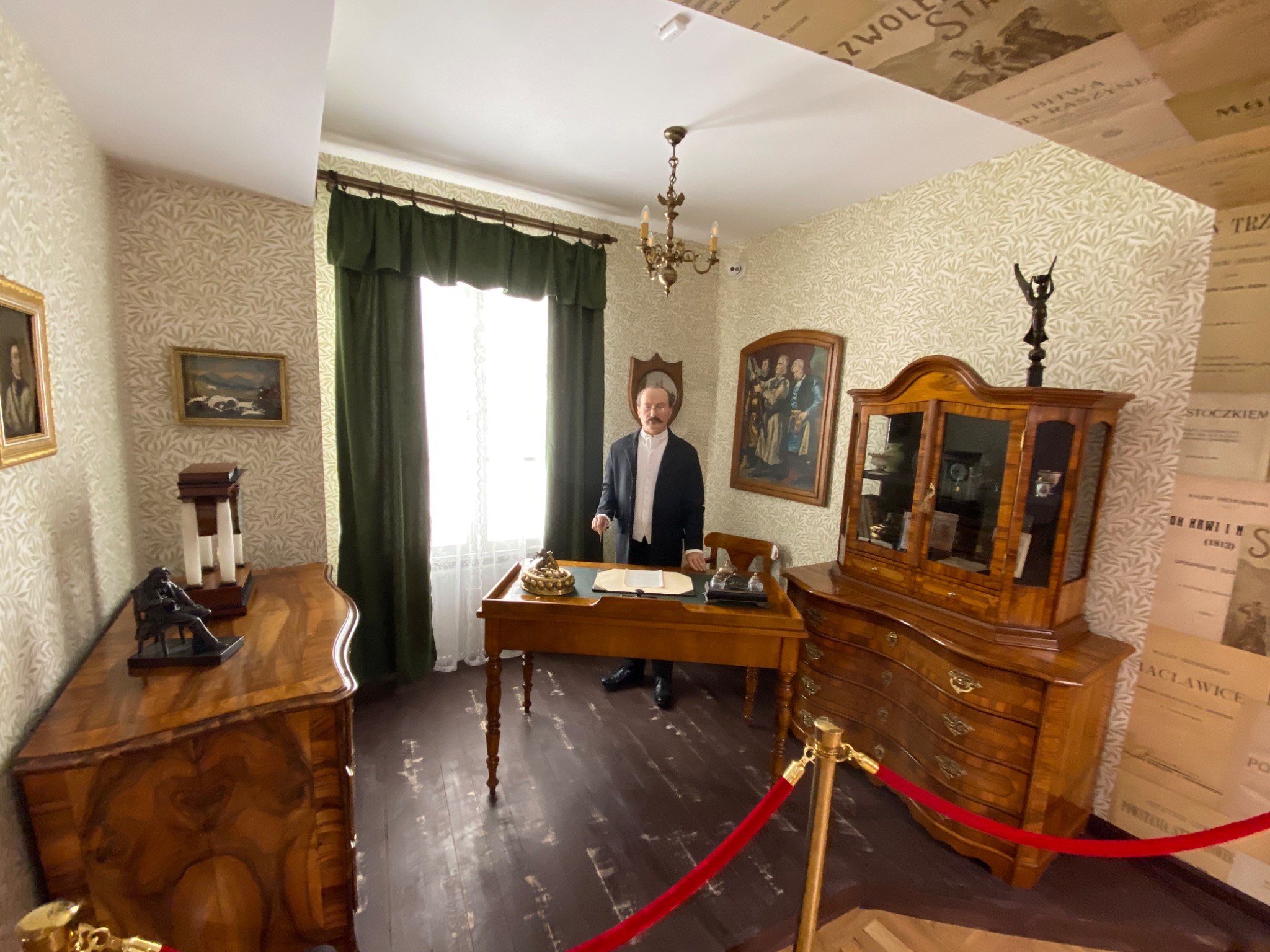 Wnętrze pokoju stylizowanego na gabinet z epoki, z drewnianymi meblami, tapetą w roślinny wzór i figurą mężczyzny w stroju z epoki przy biurku.