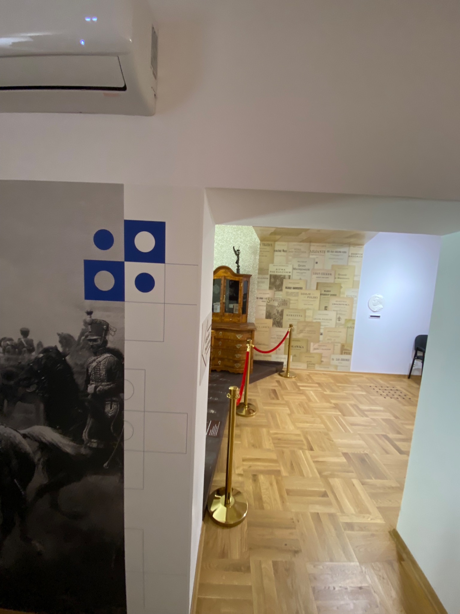 Wnętrze muzeum z parkietem, ścianą z grafiką historyczną i ekspozycją dokumentów za sznurami.