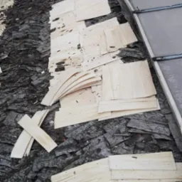 Fragment dachu z częściowo ułożonymi drewnianymi gontami, widoczne surowe deseczki i fragment starego poszycia dachowego z ciemnego materiału bitumicznego.