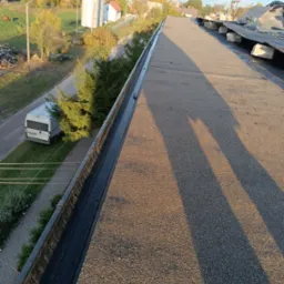 Widok z g&oacute;ry na płaski dach pokryty papą, z widoczną rynną i otoczeniem w tle, w tym budynki i drzewa w jesiennej scenerii.