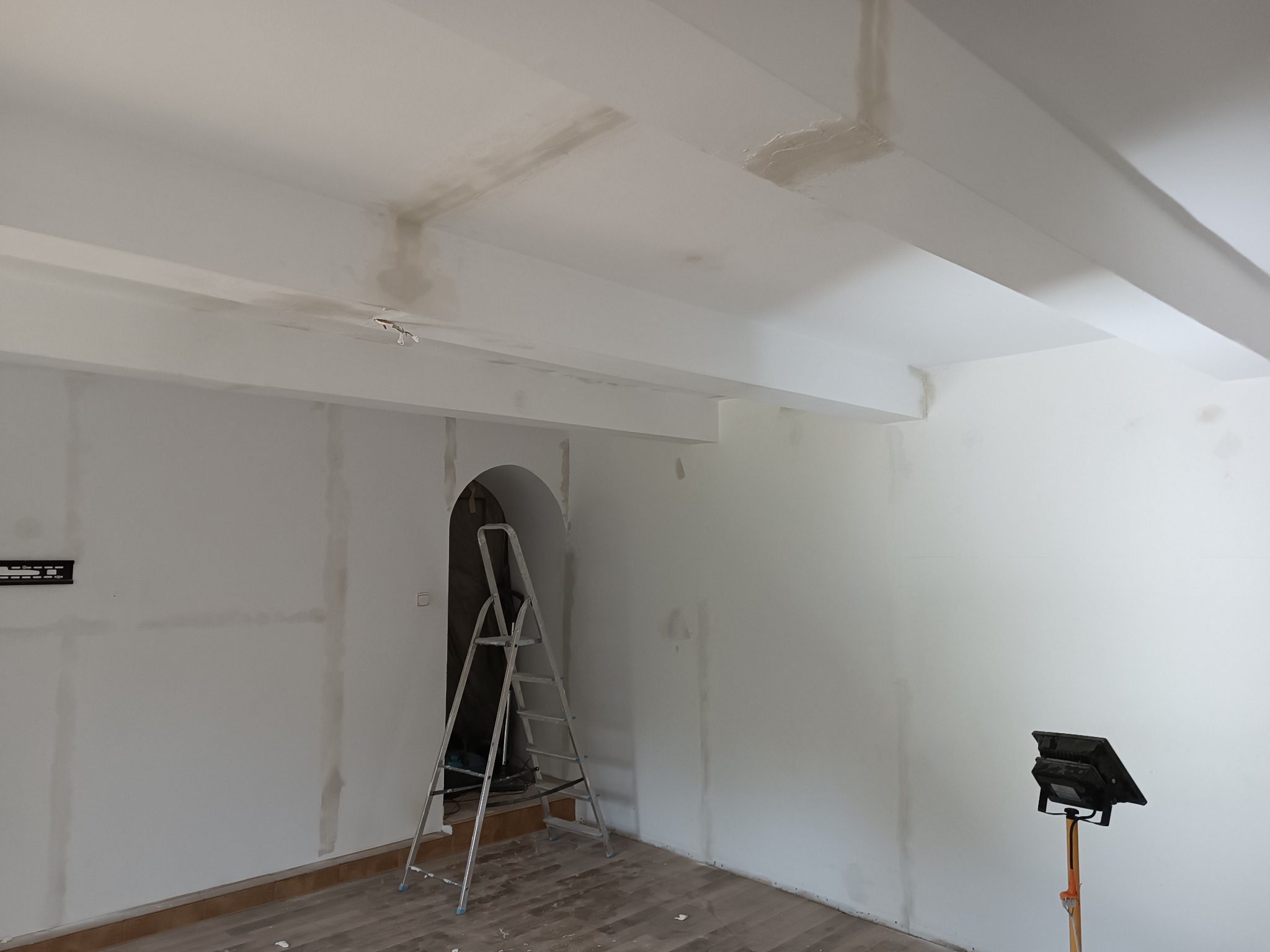 Wnętrze w trakcie prac wykończeniowych: ściany częściowo pokryte gładzią, widoczna drabina aluminiowa przy wejściu z łukiem oraz lampa budowlana na statywie. Na suficie widoczne niedoskonałości...
