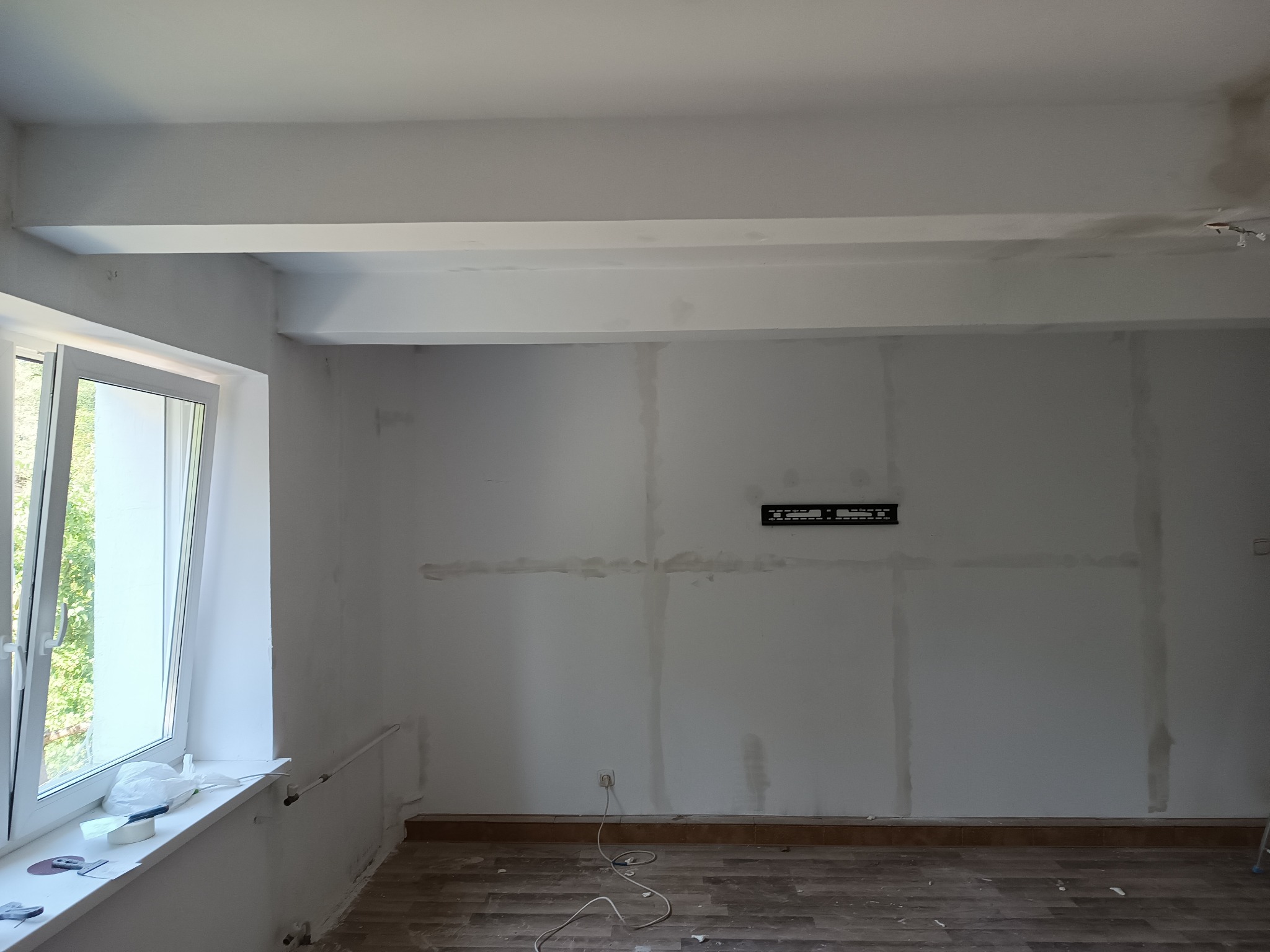 Pokój w trakcie przygotowania do malowania: widoczne ślady po szpachlowaniu na ścianach, niezagruntowane powierzchnie, listwy przypodłogowe i częściowo zabezpieczone okno. Na parapecie leżą...