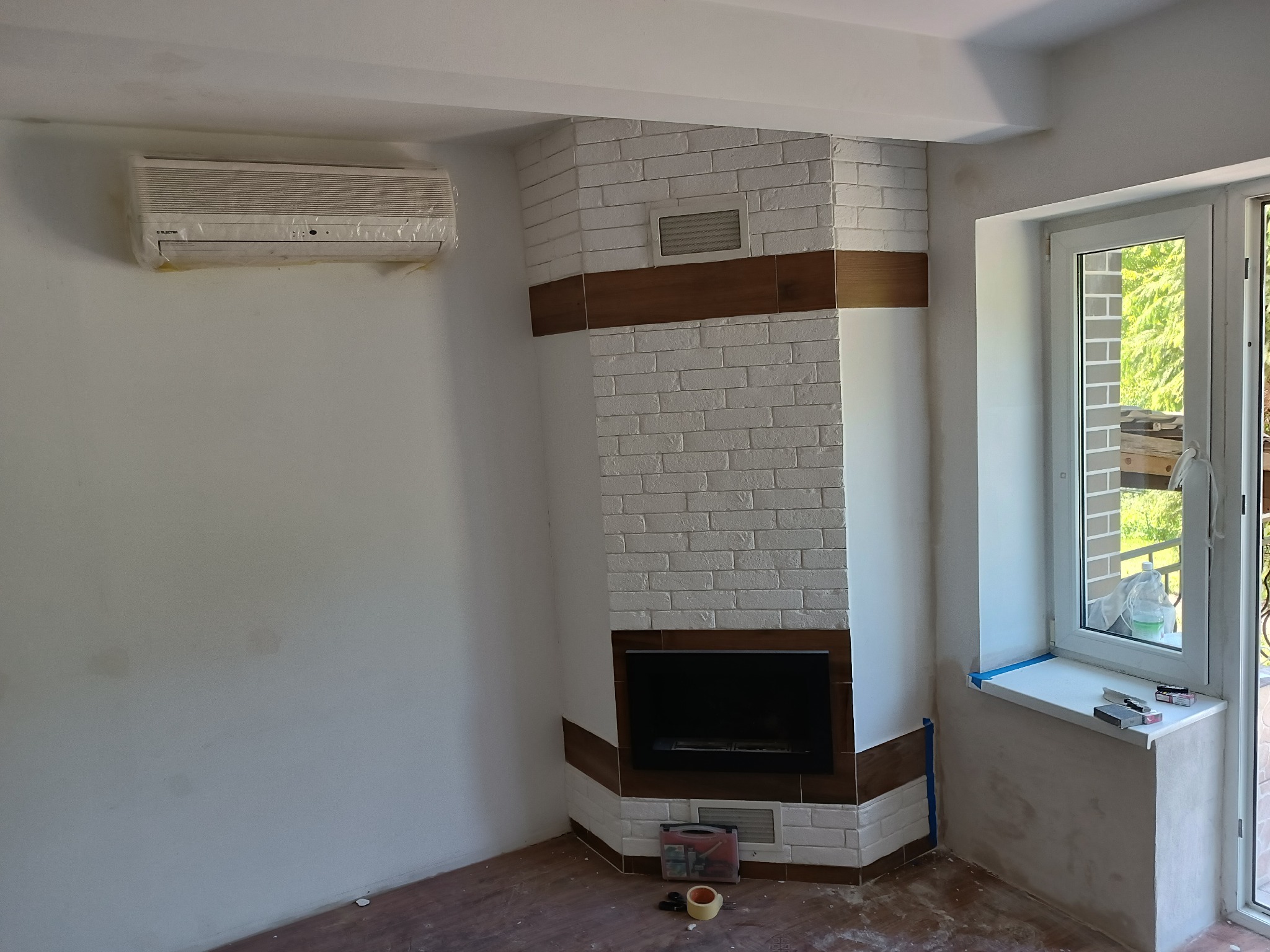 Wnętrze pokoju z wykończonym kominkiem obłożonym białą cegłą z brązowymi akcentami, klimatyzator na ścianie w folii ochronnej, okno z widokiem na zieleń, oraz narzędzia i akcesoria na podłodze...