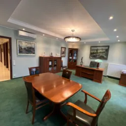 Przestronne biuro z drewnianym stołem konferencyjnym, zielonym dywanem, biurkiem z lampką, regałami z książkami i obrazami na ścianach, oświetlone lampą sufitową.