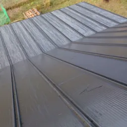 Nowoczesne, grafitowe pokrycie dachowe z blachy na ukończonym dachu, widoczne krople wody po deszczu, w tle trawa i drewniane palety.