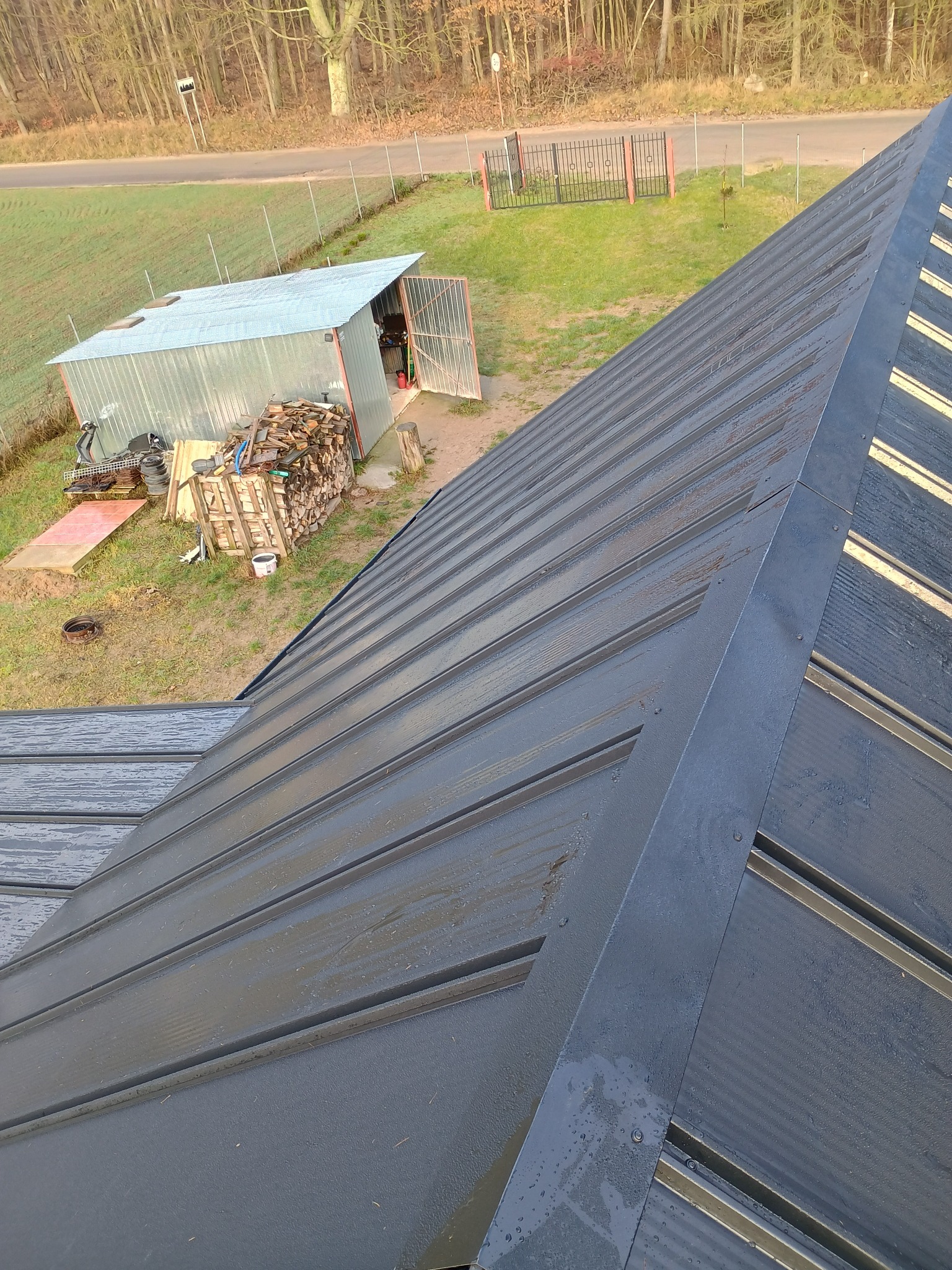 Nowe, grafitowe pokrycie dachowe z blachy trapezowej, widok z góry, w tle budynek gospodarczy i stos drewna.