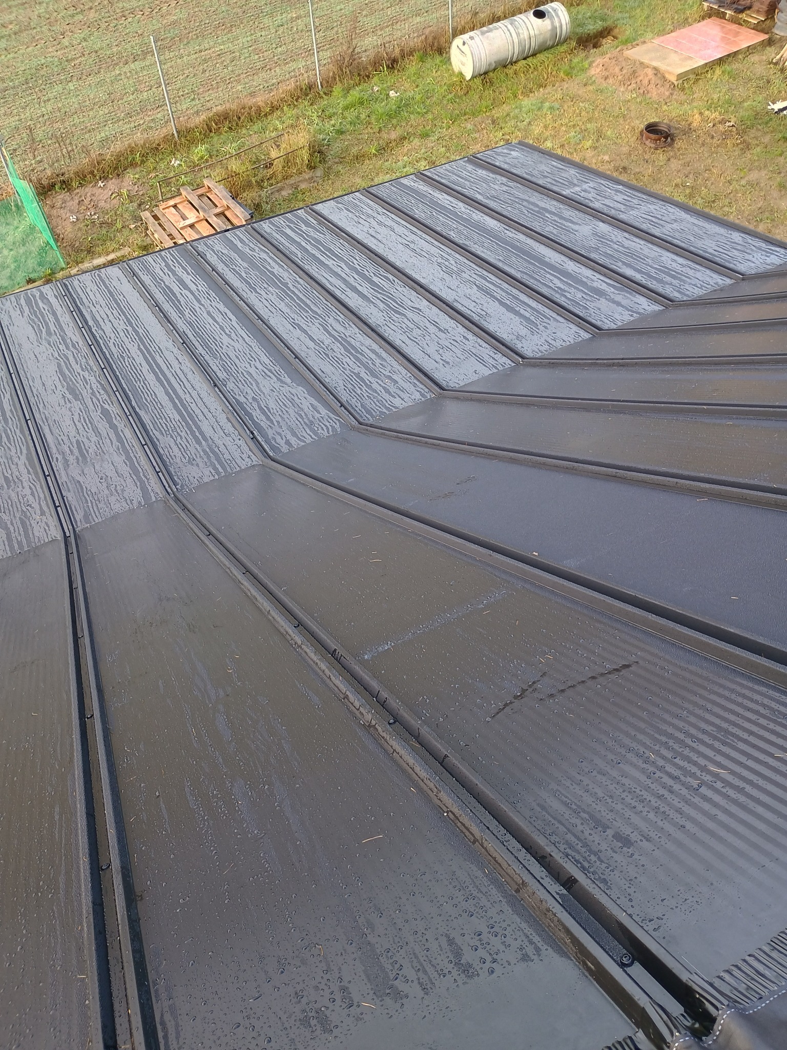Nowoczesne, grafitowe pokrycie dachowe z blachy na ukończonym dachu, widoczne krople wody po deszczu, w tle trawa i drewniane palety.