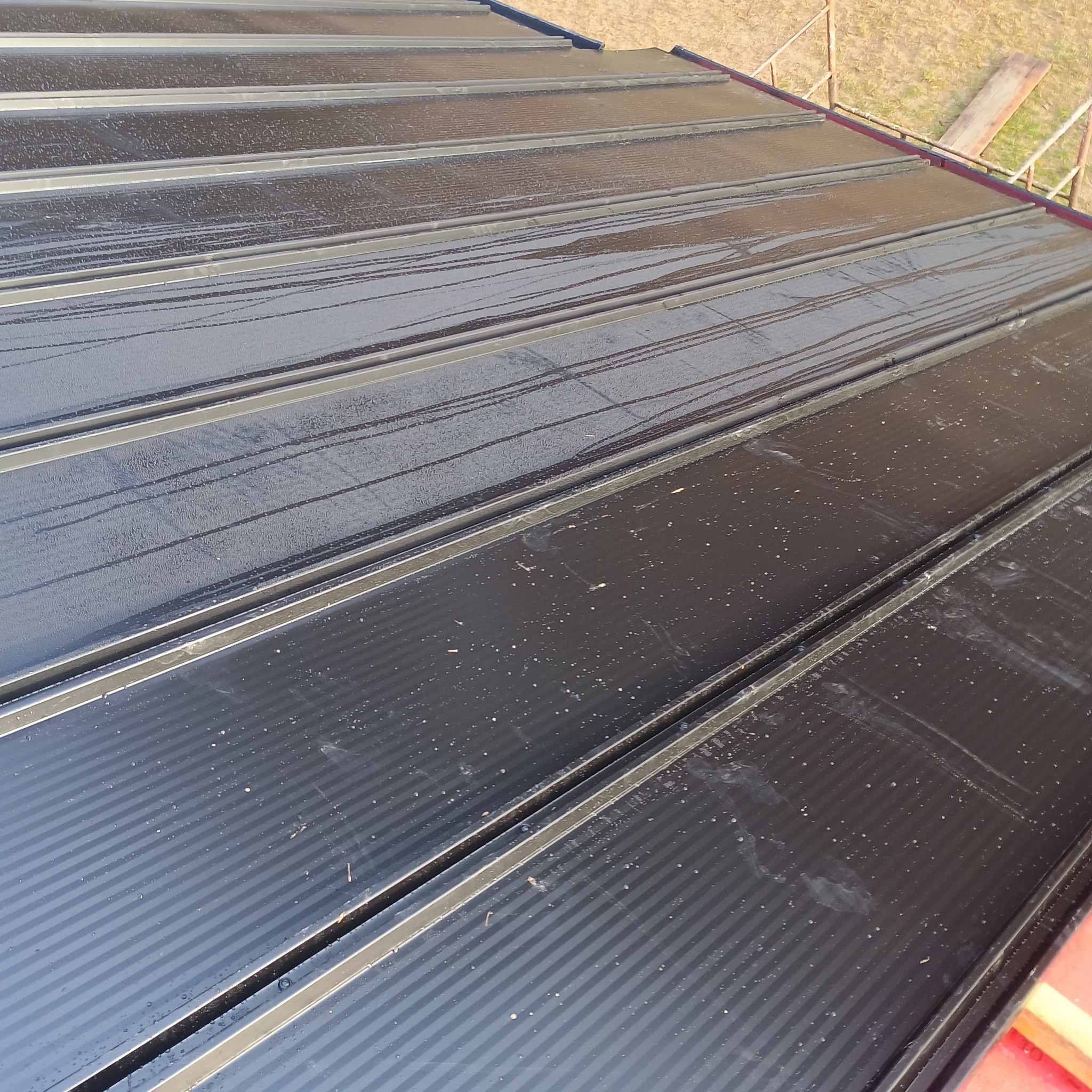 Błyszczące, czarne blaszane pokrycie dachowe z widocznymi śladami wody i lekkimi zabrudzeniami, ułożone na konstrukcji dachu widocznej w tle.