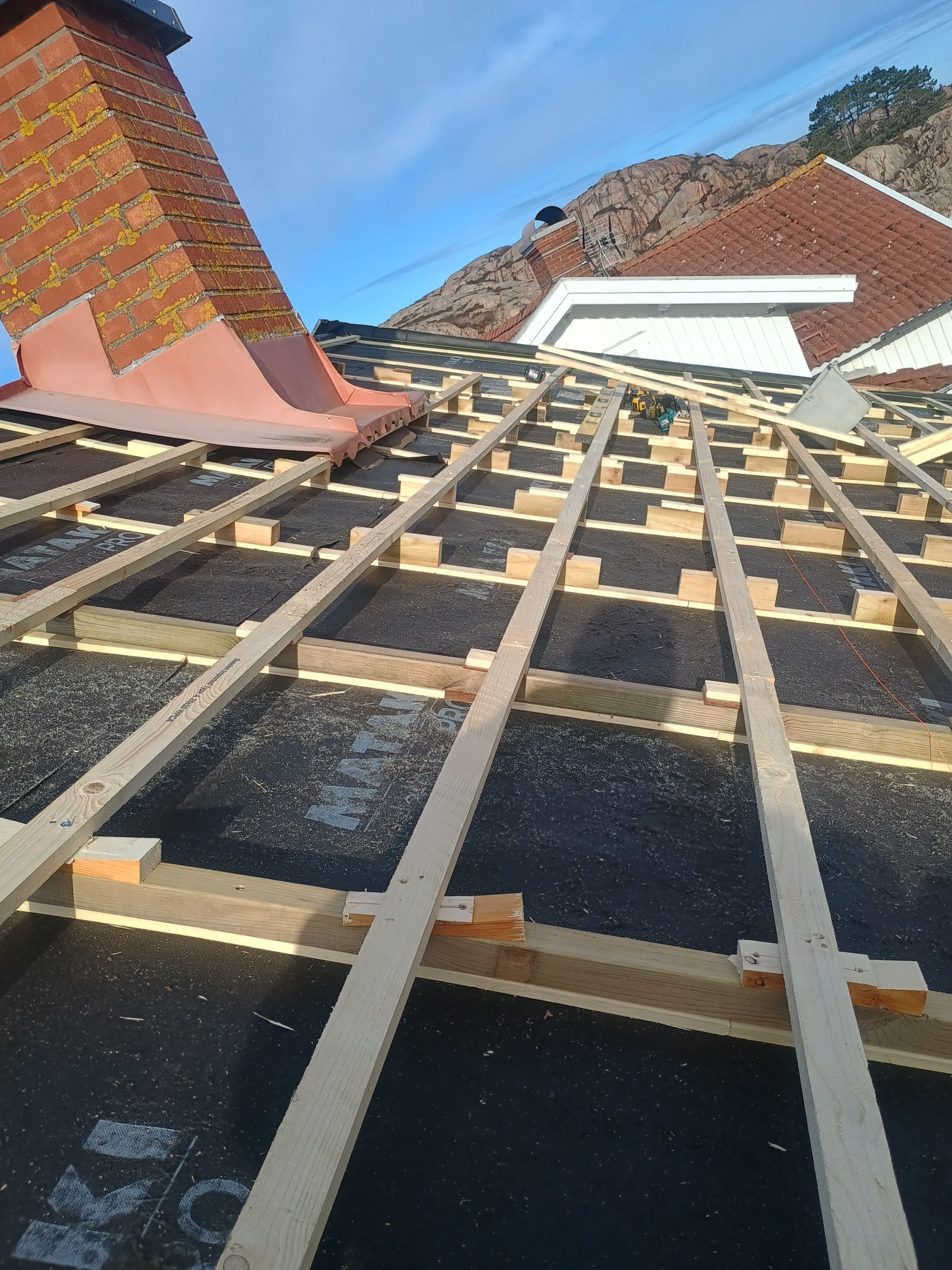 Konstrukcja drewniana dachu w trakcie budowy, widoczne deski, membrana i komin z cegły na tle nieba i wzgórz.