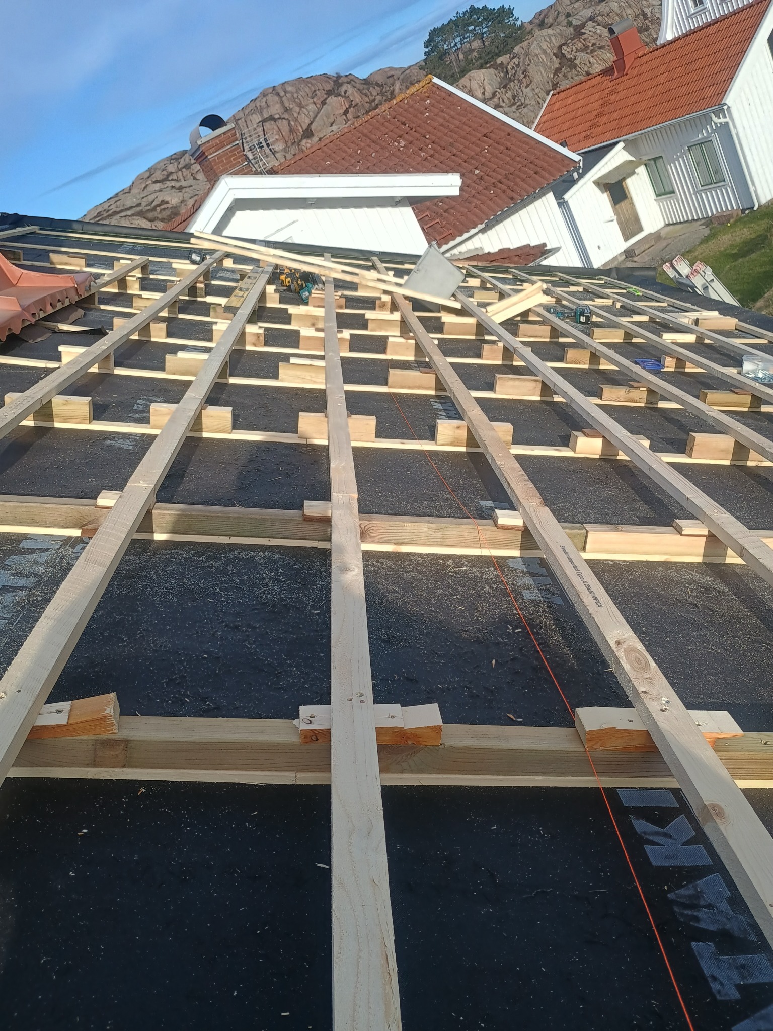Konstrukcja drewniana dachu w trakcie budowy, widoczne łaty i kontrłaty na czarnej membranie dachowej, w tle domy i skały.