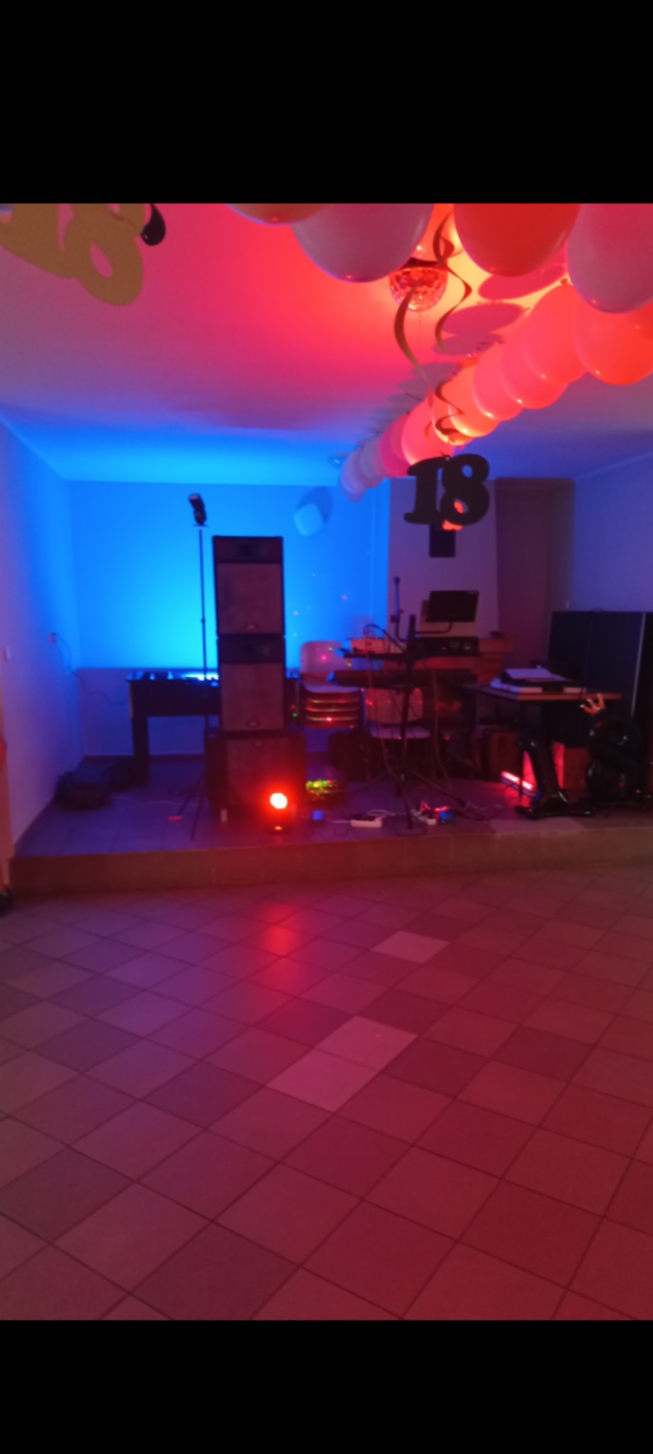 Sala z podwyższeniem, na którym znajduje się sprzęt muzyczny, w tle dekoracje urodzinowe, oświetlenie sceniczne w kolorach czerwonym i niebieskim.