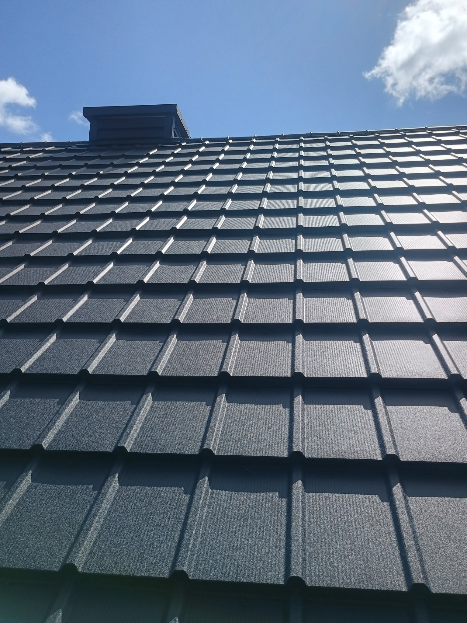 Grafitowy dach z blachodachówki modułowej w zbliżeniu, widoczny kominek wentylacyjny i błękitne niebo z chmurami w tle. Nowoczesne pokrycie dachowe.