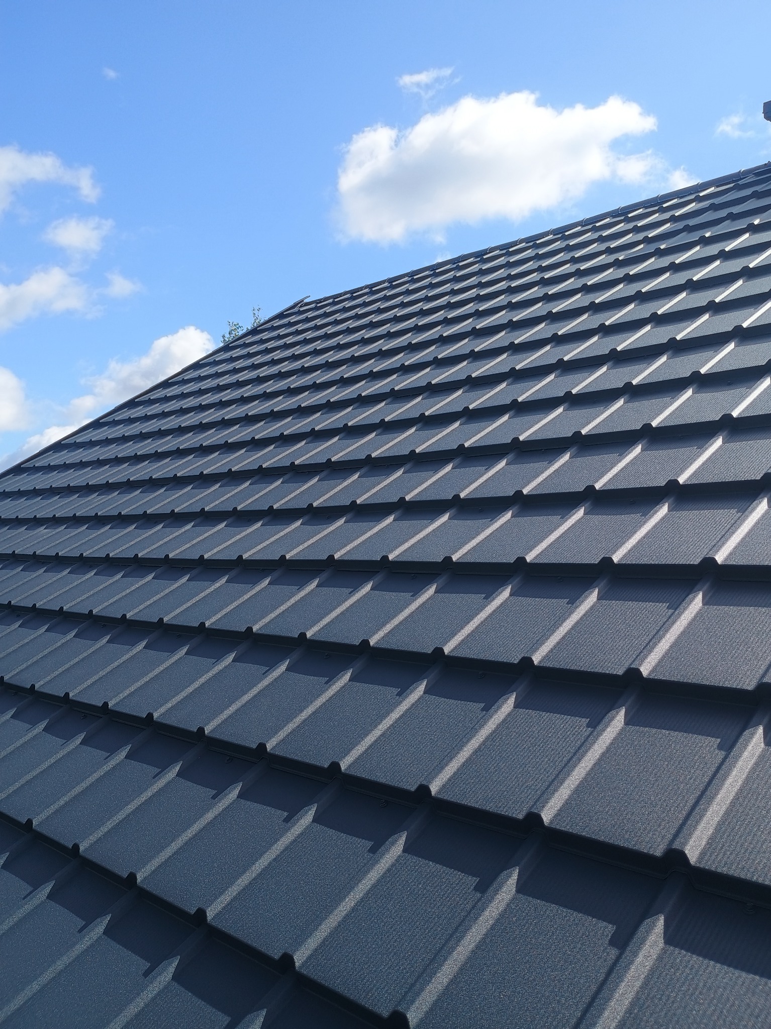 Nowoczesne krycie dachu z ciemnej blachy trapezowej na tle błękitnego nieba z delikatnymi obłokami. Widok z dołu, podkreślający regularność i estetykę wykonania.