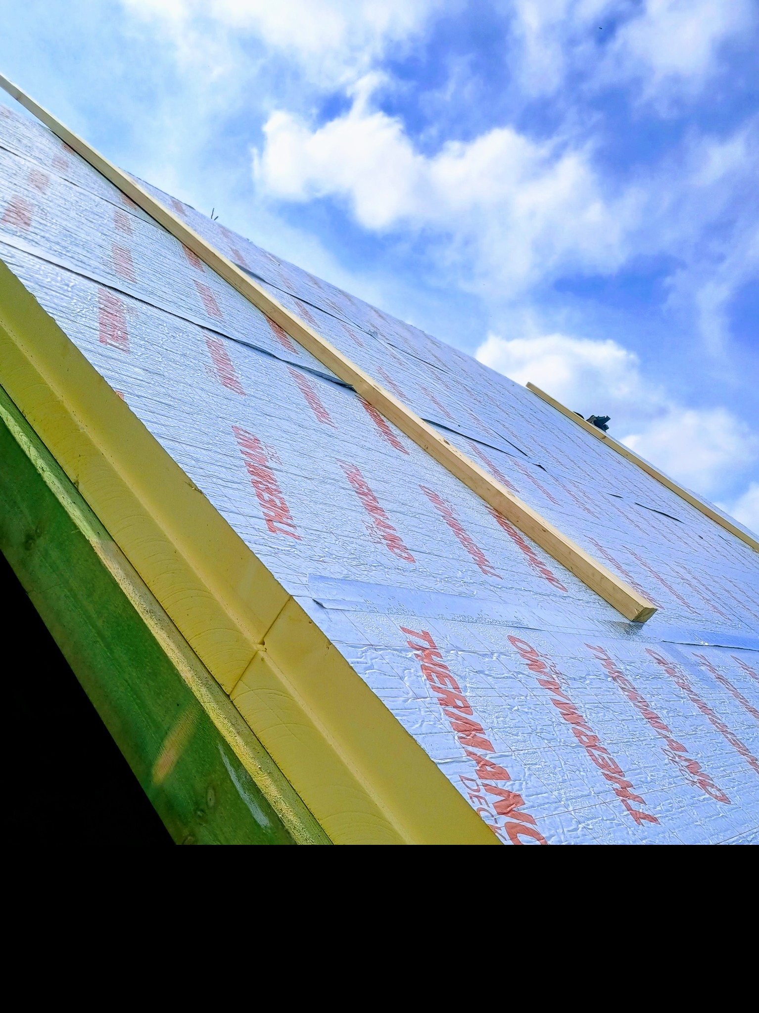 Ocieplenie dachu folią termoizolacyjną z widocznymi łatami drewnianymi na tle błękitnego nieba z chmurami. Ujęcie z dołu, ukazujące detale montażu.