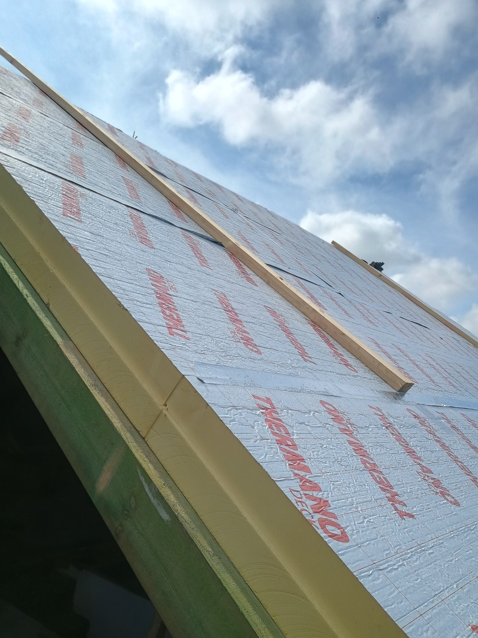 Dach z izolacją Thermano, widok pod kątem. Elementy drewniane konstrukcji dachu, niebieskie niebo z chmurami w tle. Detal izolacji dachu podczas montażu.