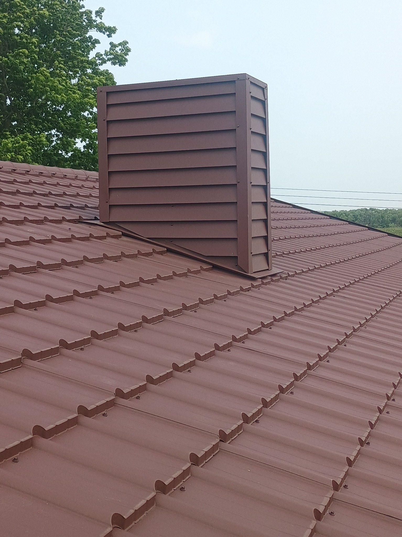 Ciemnobrązowy dach z dachówki ceramicznej z kominem w tym samym kolorze, widok z góry, na tle zieleni drzew i jasnego nieba.