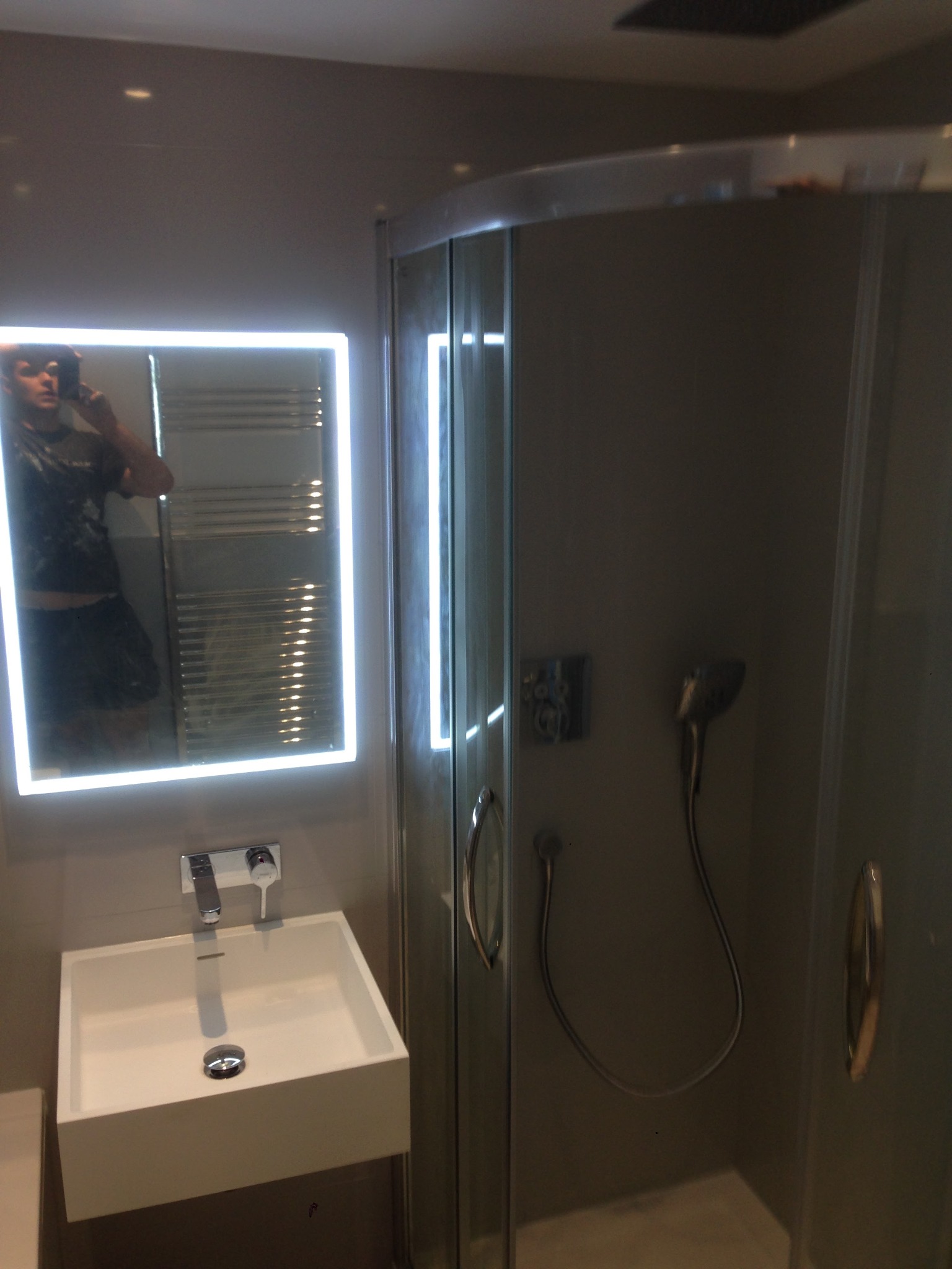 Nowoczesna łazienka z kabiną prysznicową, prostokątną umywalką i podświetlanym lustrem, w którym odbija się osoba robiąca zdjęcie.