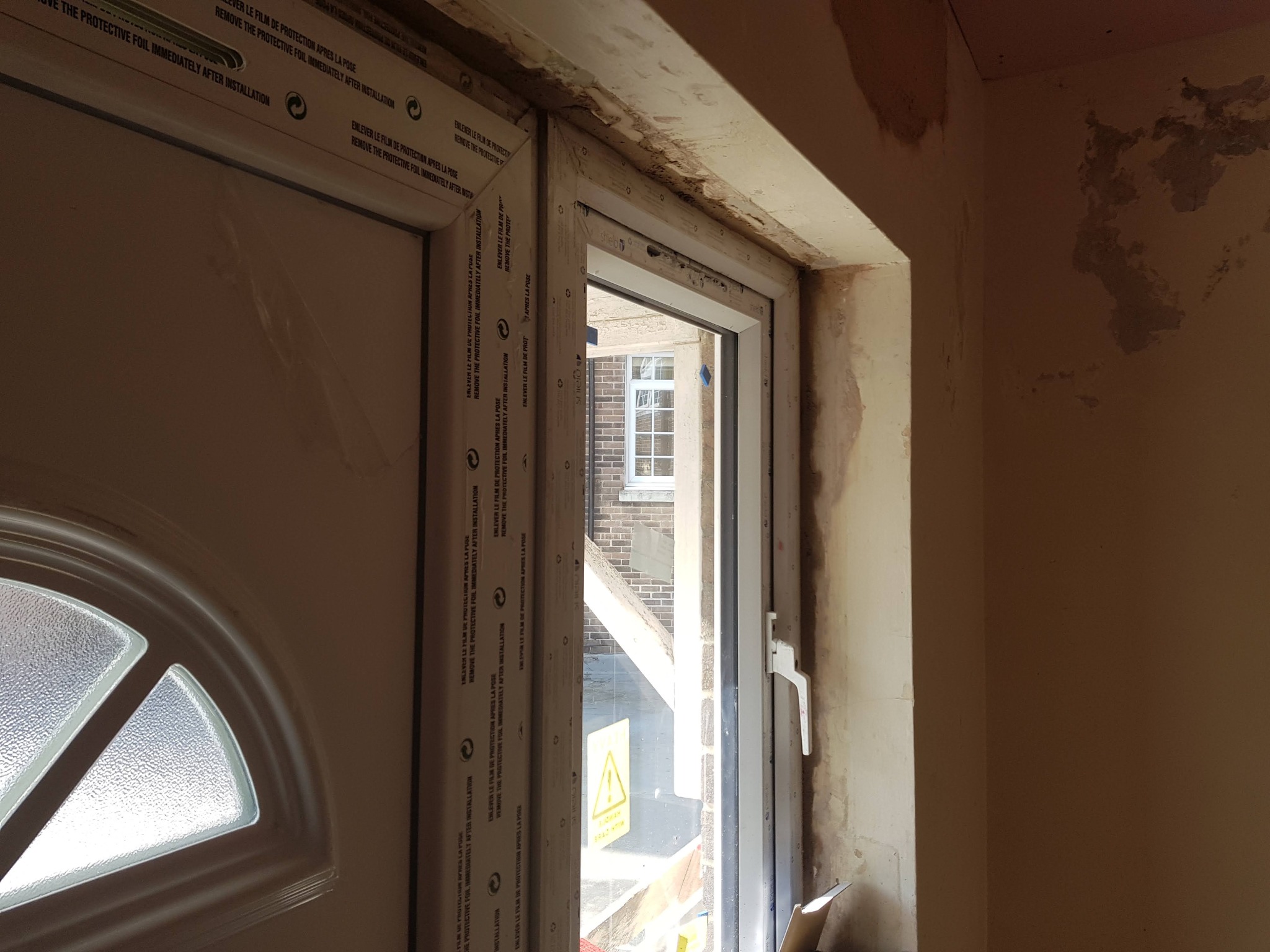 Nowe, białe drzwi wejściowe z szybą w kształcie półkola, jeszcze z folią ochronną, osadzone w surowym otworze, ściany wymagają wykończenia.