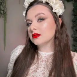 Portret kobiety z kwiatowym wiankiem, intensywnym makijażem ust i oczu, na tle dekoracji roślinnych. Uwagę przyciąga heterochromia.