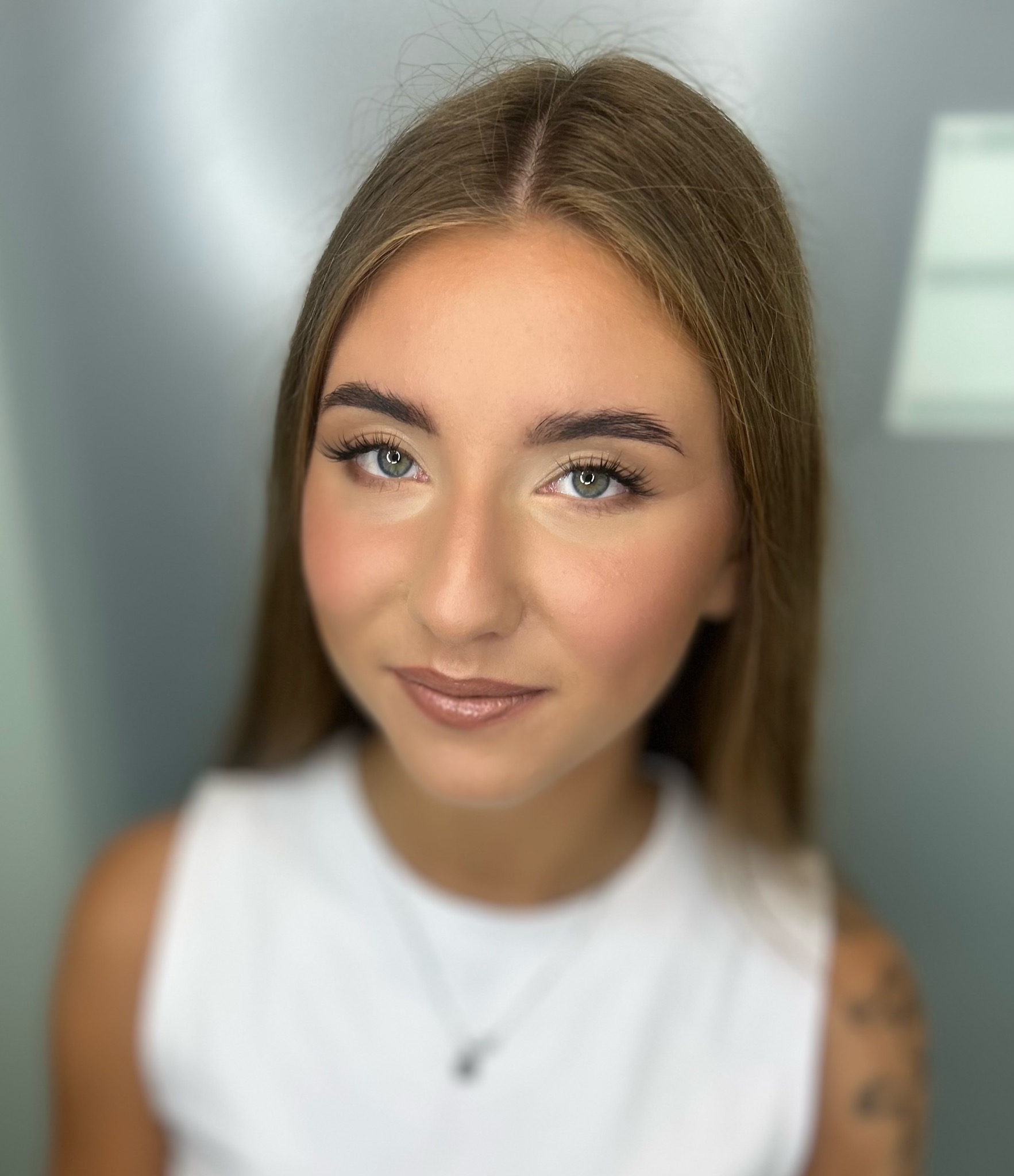 Portret młodej kobiety z delikatnym, naturalnym makijażem, podkreślonymi brwiami i błyszczącymi ustami, na rozmytym tle w chłodnych odcieniach szarości.