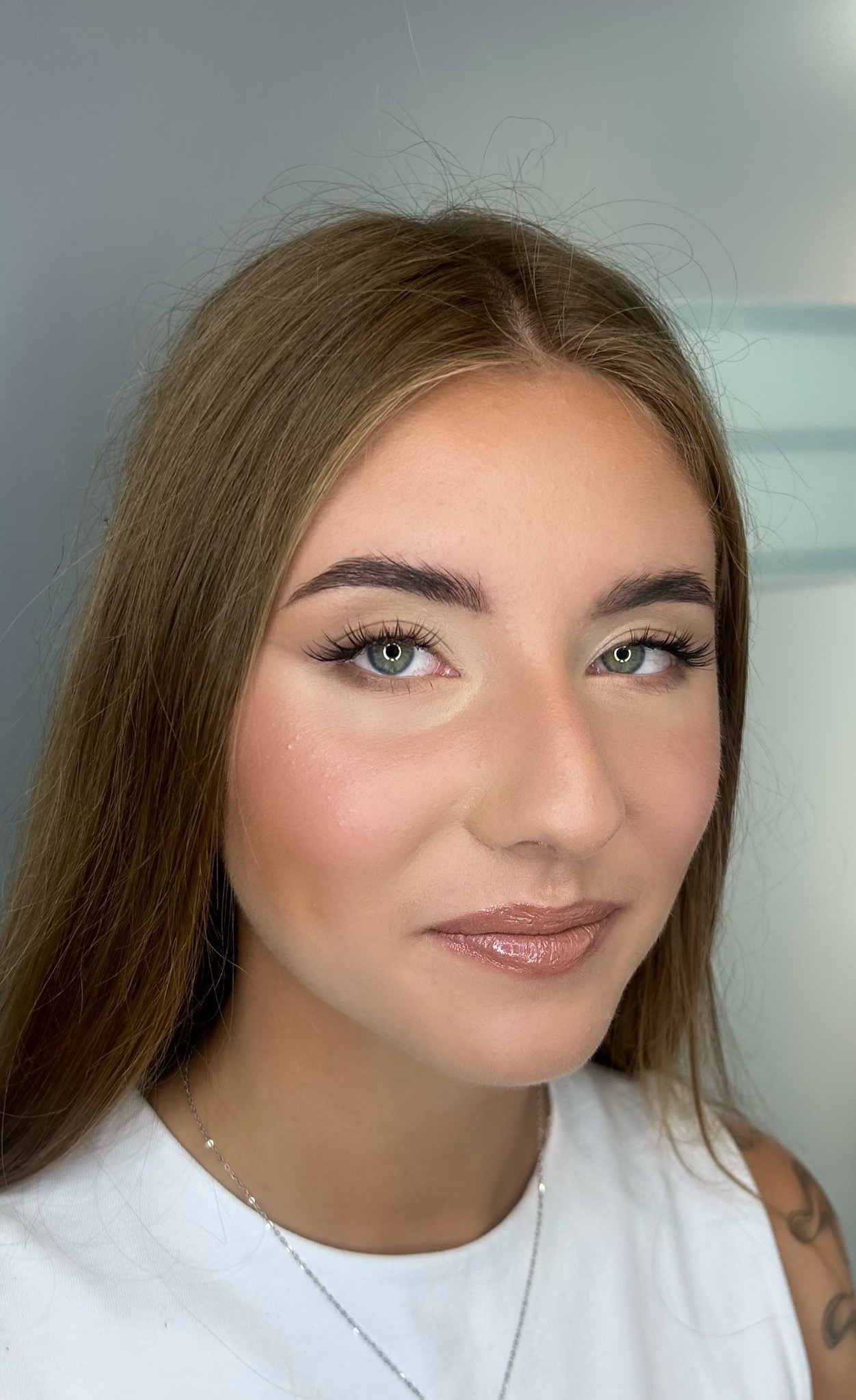Portret młodej kobiety z delikatnym, dziennym wizażem: podkreślone oczy z sztucznymi rzęsami, brwi o naturalnym kształcie, rozświetlona cera z subtelnym różem i błyszczące usta w odcieniu nude.