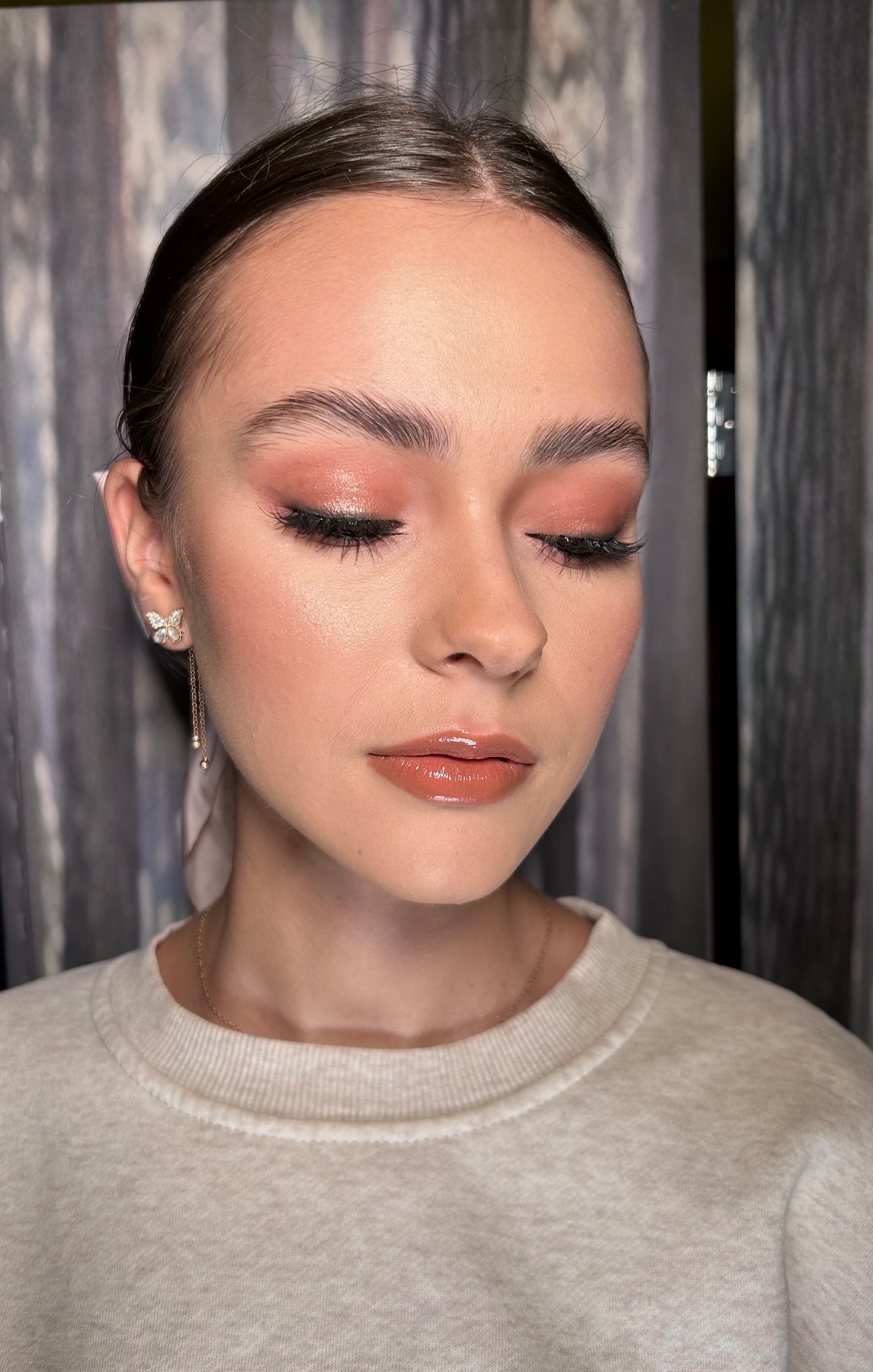 Portret kobiety z zamkniętymi oczami prezentującej delikatny, błyszczący, brzoskwiniowy odcień na powiekach, podkreślone rzęsy, konturowanie policzków oraz błyszczące usta w kolorze nude,...