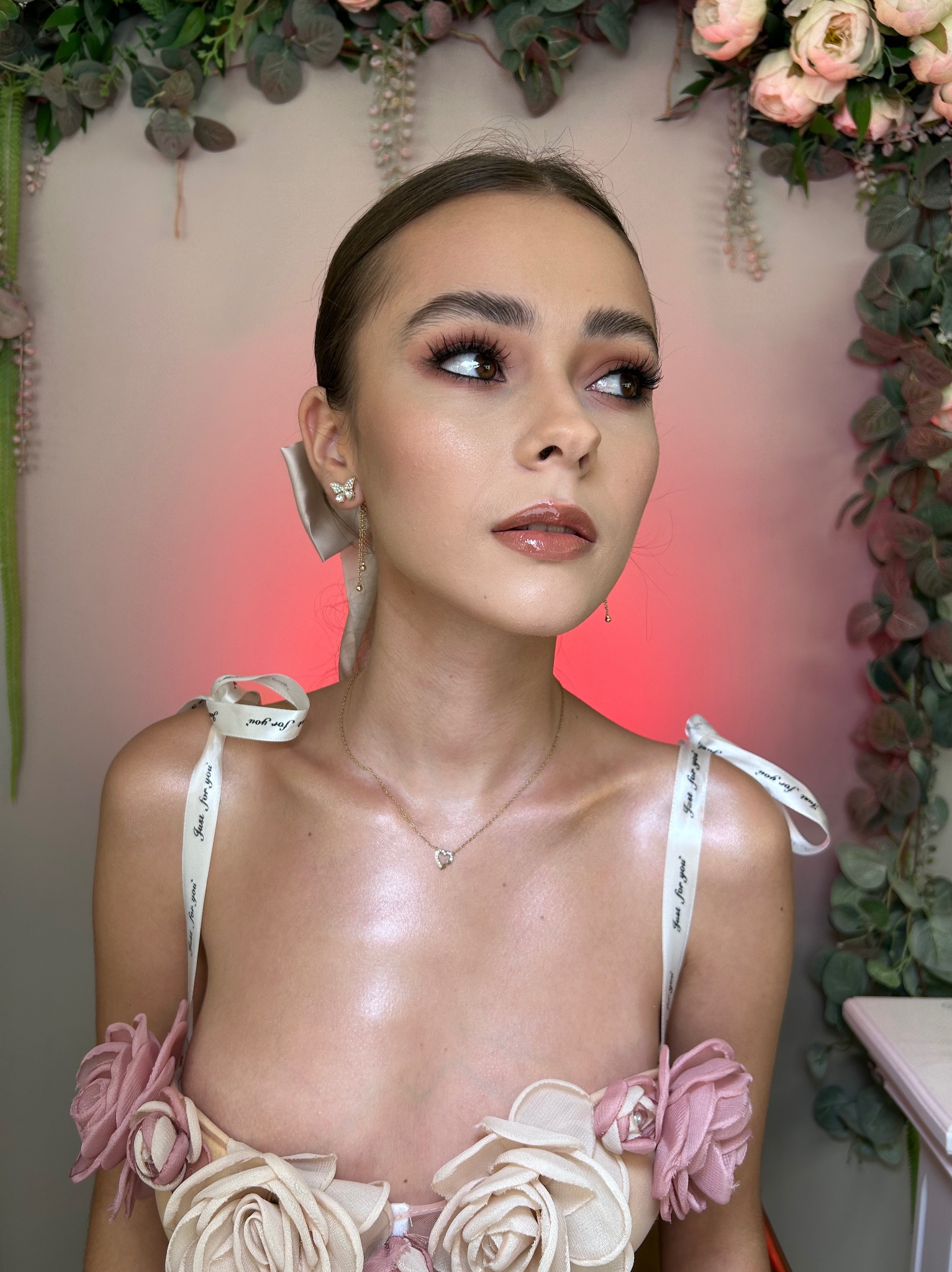 Portret kobiety z profesjonalnym makijażem oczu i ust w odcieniach brązu, z upiętymi włosami z kokardą, złotymi kolczykami w kształcie motylków i naszyjnikiem z serduszkiem, na tle ściany...