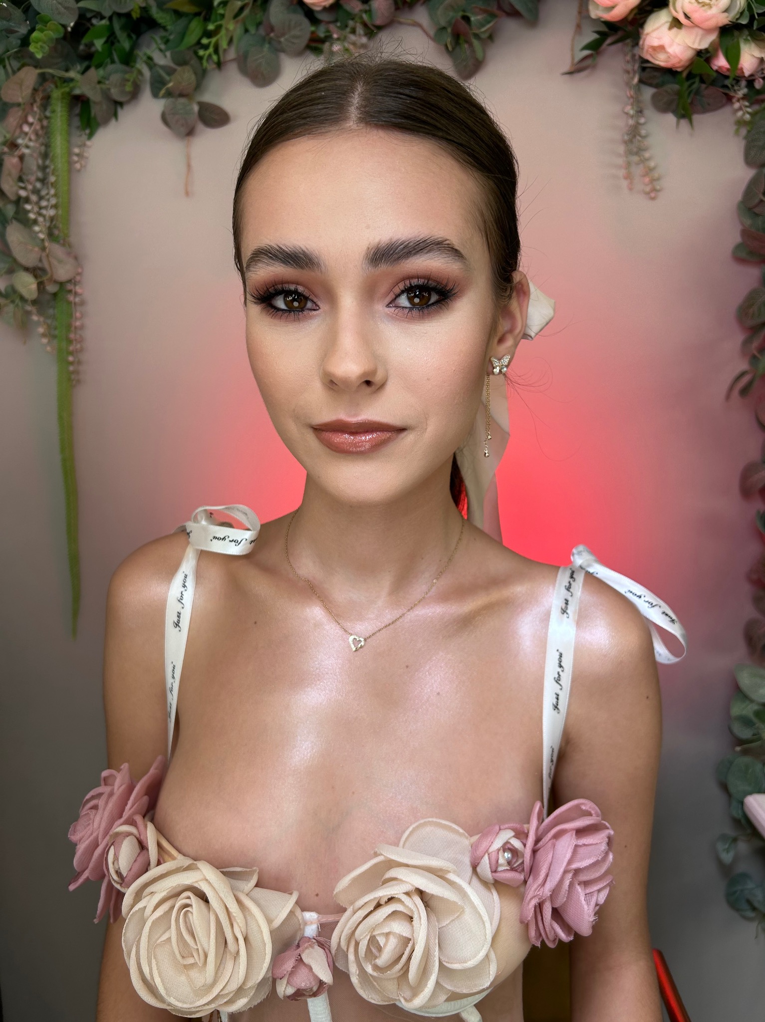 Portret młodej kobiety z profesjonalnym, błyszczącym makijażem oczu w odcieniach brązu i błyszczykiem, włosy upięte w kokardę, biżuteria w postaci kolczyków motylków i naszyjnika z sercem, ozdobny...