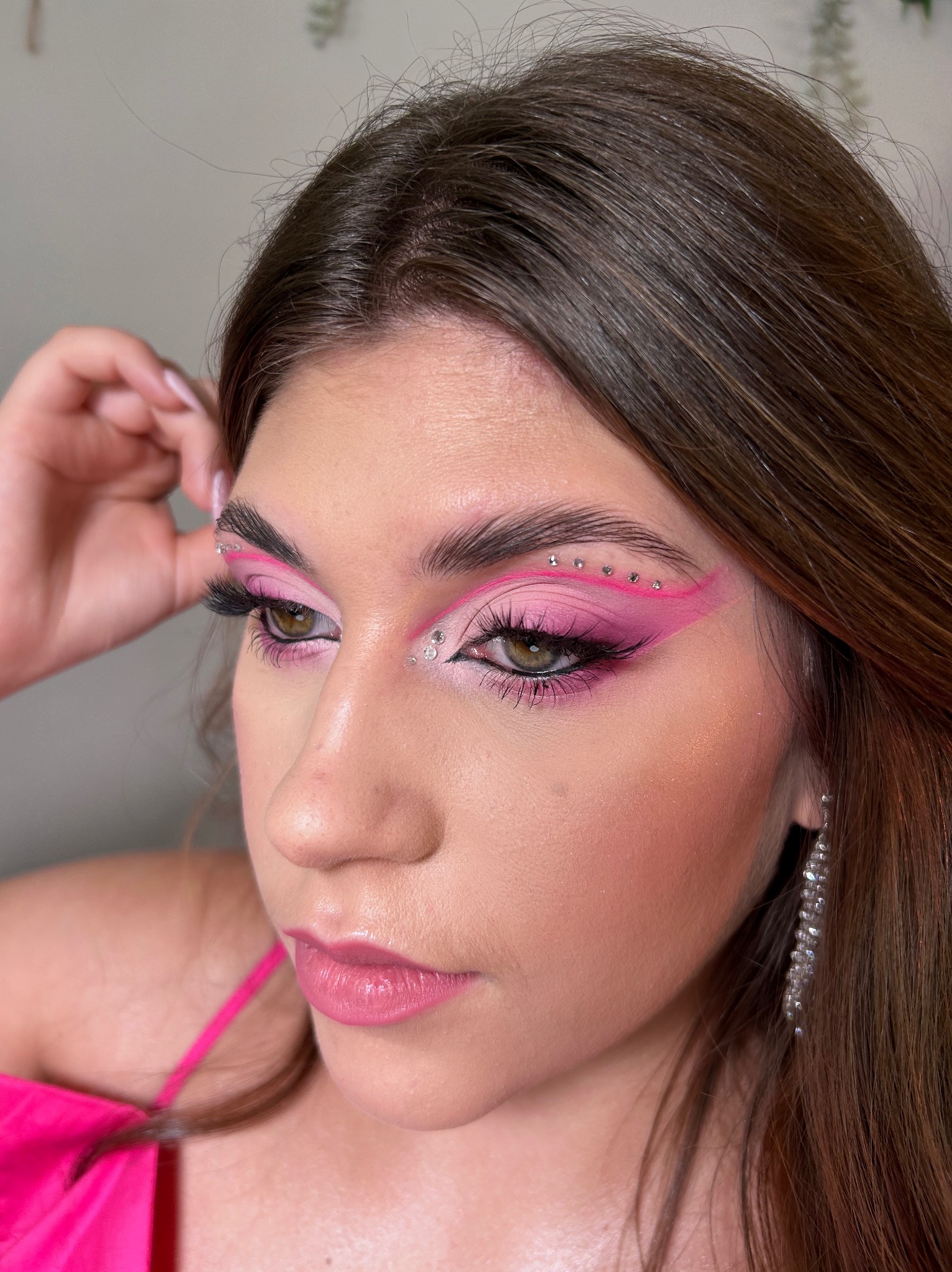 Portret kobiety z intensywnym, różowym makijażem oczu z geometrycznymi liniami i błyszczącymi kryształkami, uzupełnionym o błyszczące usta i długie kolczyki.