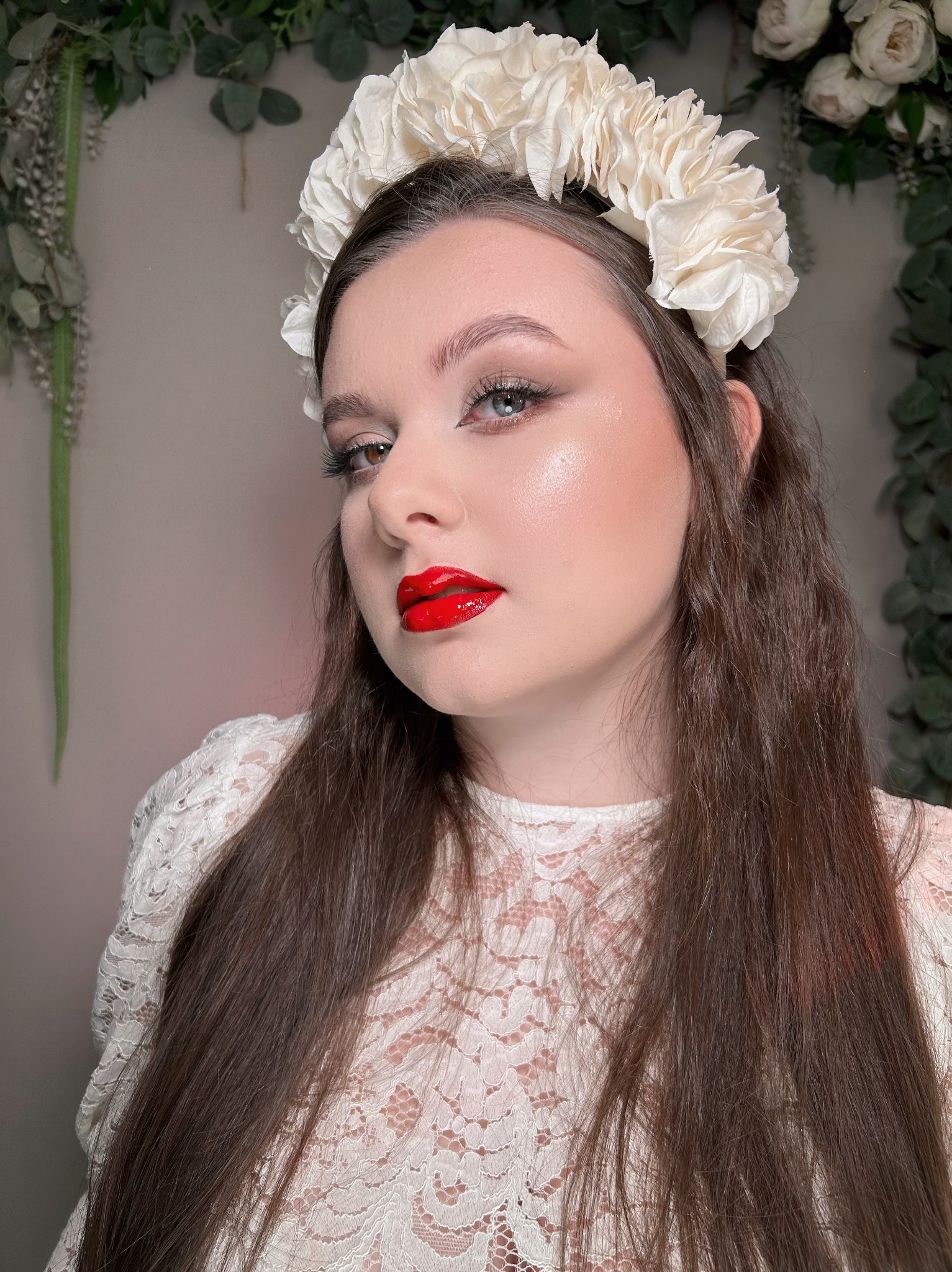 Portret kobiety z kwiatowym wiankiem, intensywnym makijażem ust i oczu, na tle dekoracji roślinnych. Uwagę przyciąga heterochromia.