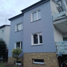 Remonty ogólnobudowlane - Dwukondygnacyjny dom z niebieską elewacją i kamienną podmurówką, widoczne rusztowanie na balkonie i fragment roweru, trawnik z małym drzewkiem.