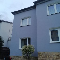 Remonty ogólnobudowlane - Dwupiętrowy dom z niebieską elewacją i kamienną podmurówką, z pracownikiem w trakcie czyszczenia lub przygotowywania terenu wokół budynku, widoczne okna z białymi ramami i niewielkie drzewo przed...