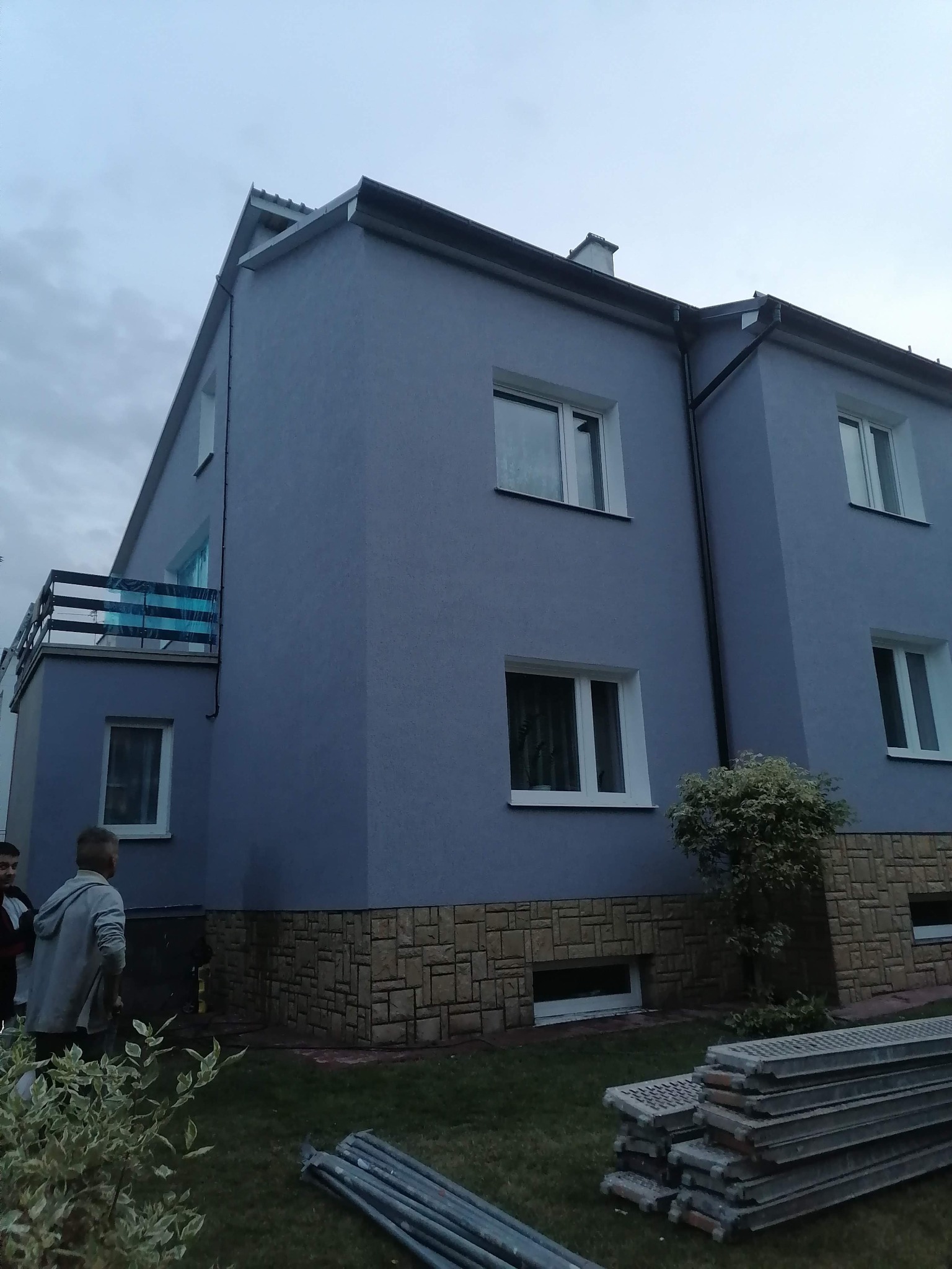 Dwupiętrowy budynek z szarą elewacją, wykończony kamieniem na dole, z widocznymi rusztowaniami i osobami w pobliżu, prawdopodobnie po świeżo zakończonym malowaniu.