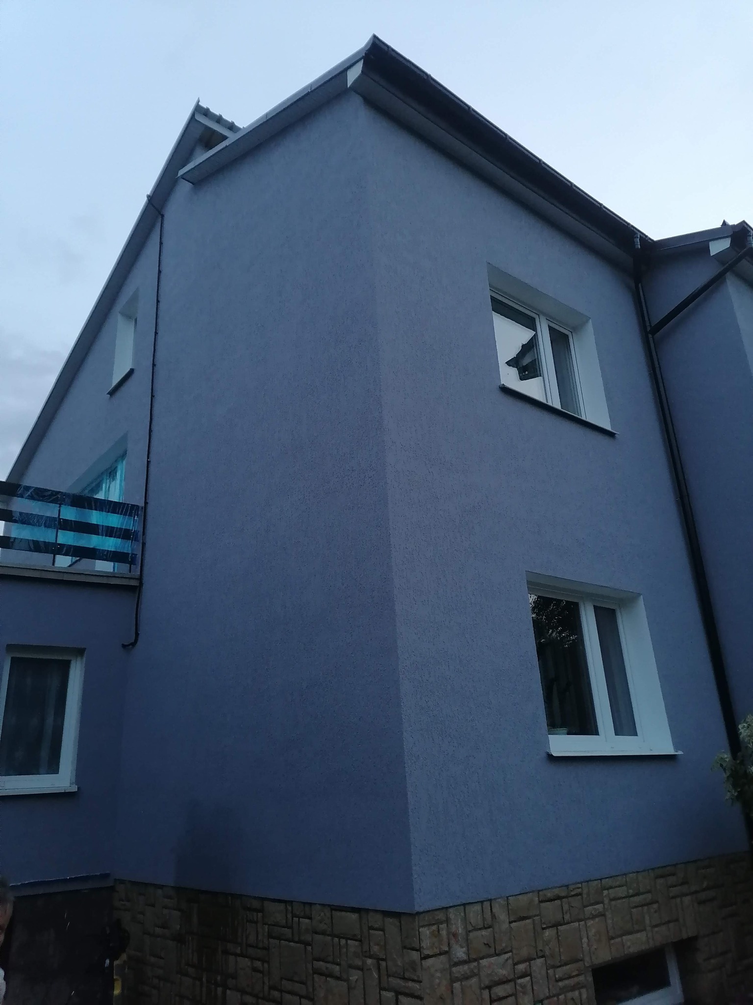 Dwupiętrowy dom z szarą elewacją i kamienną podmurówką, widok z dołu, widoczne okna z białymi ramami i balkon z niebieską zasłoną.