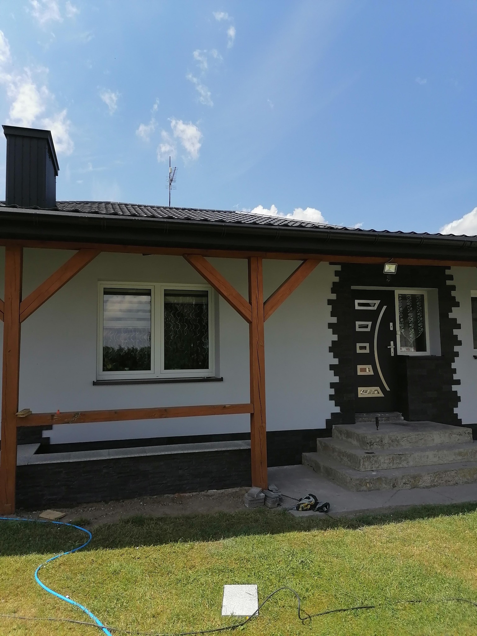 Dom jednorodzinny z nową, czarno-białą elewacją z kamienia dekoracyjnego wokół wejścia, drewniana konstrukcja werandy w trakcie budowy, widoczne narzędzia na trawniku.