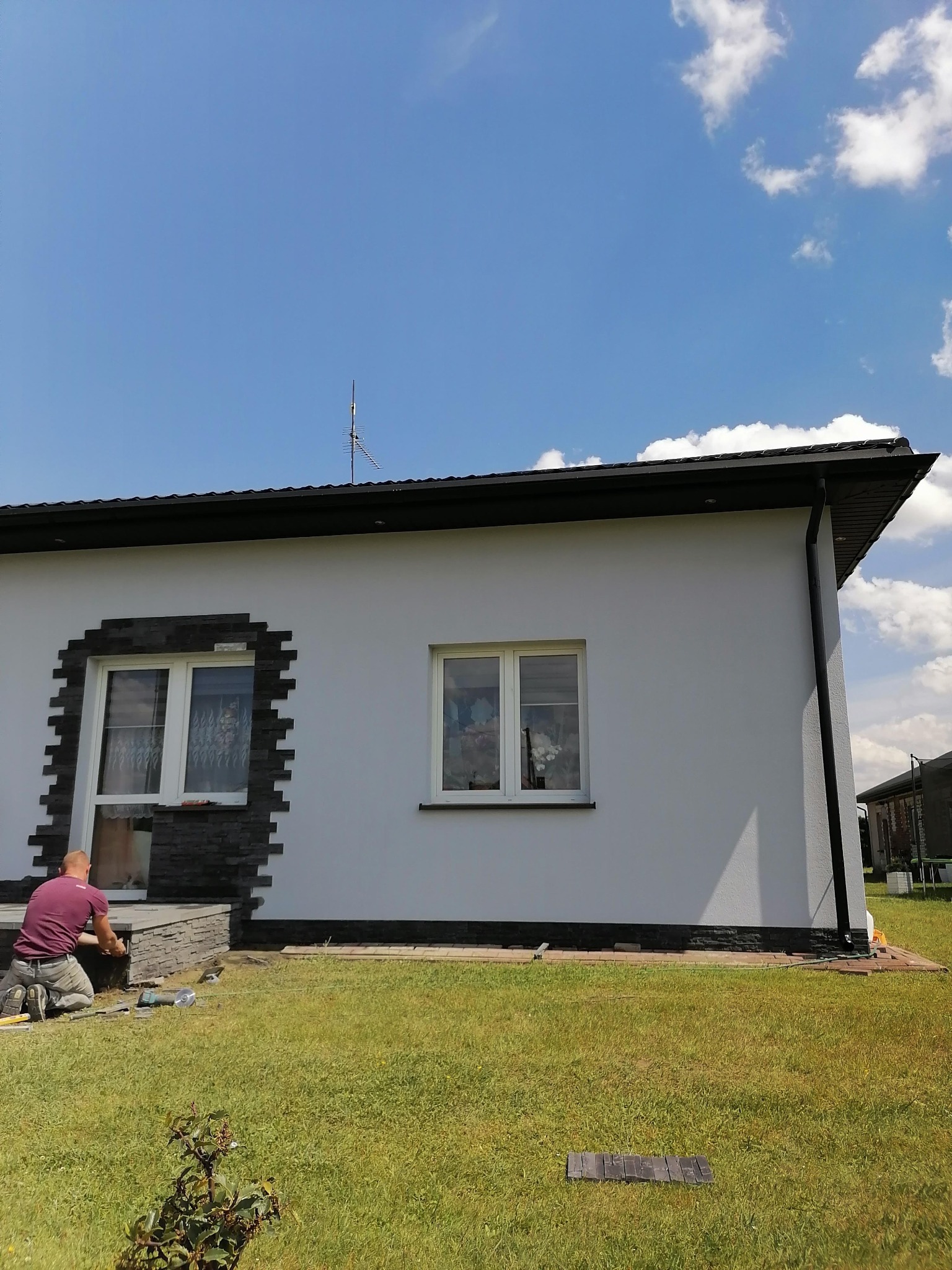 Pracownik na kolanach montuje dekoracyjną kamienną okładzinę wokół okna i na schodach wejściowych do domu z białymi ścianami i czarnym dachem.