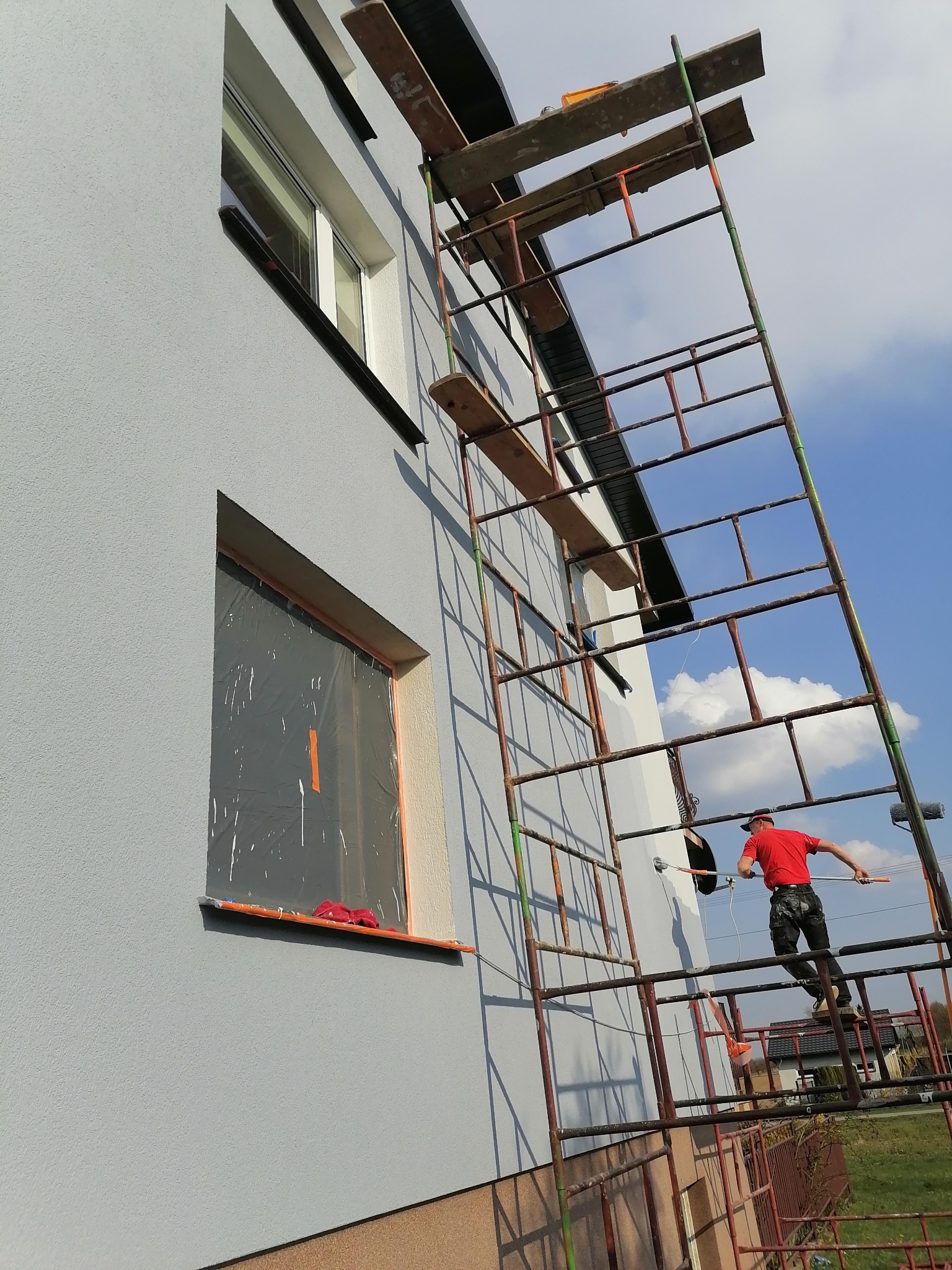 Mężczyzna w czerwonej koszulce maluje szarą elewację domu z rusztowania, okna zabezpieczone folią.