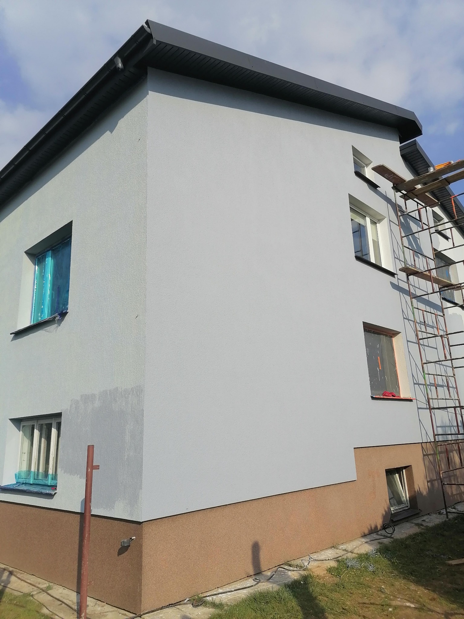 Dwukondygnacyjny dom z nową, szarą elewacją, częściowo zakryte okna folią ochronną, widoczne rusztowanie i brązowy cokół.