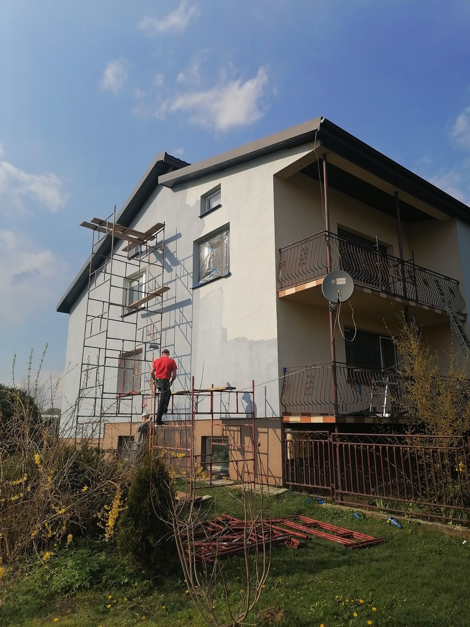 Malowanie elewacji domu jednorodzinnego ze stalowych rusztowań, widoczne fragmenty pomalowanej ściany i okna zabezpieczone folią.