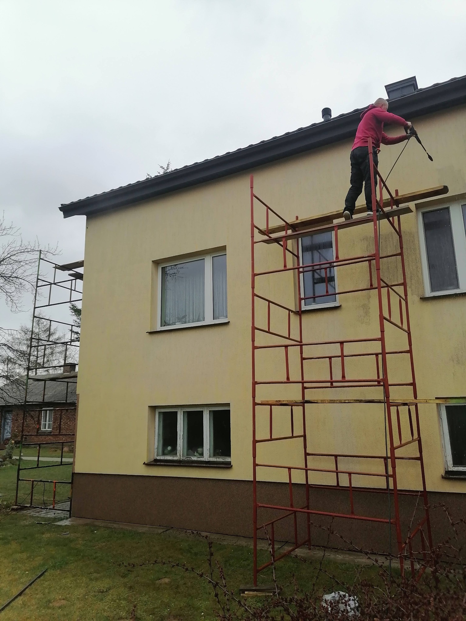 Pracownik na rusztowaniu myje elewację domu za pomocą myjki ciśnieniowej, przygotowując ją do malowania.
