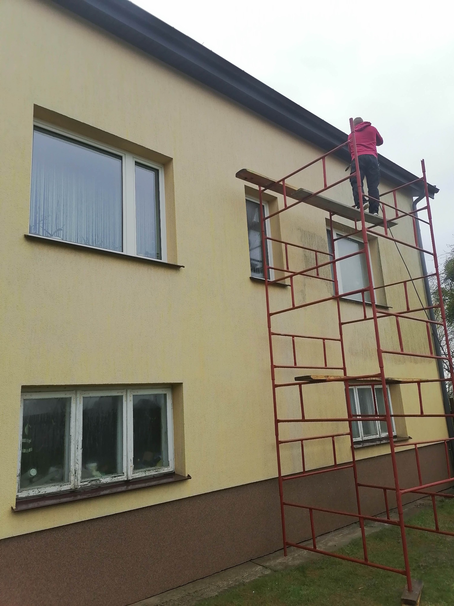 Pracownik w czerwonej bluzie na rusztowaniu maluje elewację dwupiętrowego budynku w jasnym kolorze, widoczne okna z białymi ramami i ciemna podmurówka.