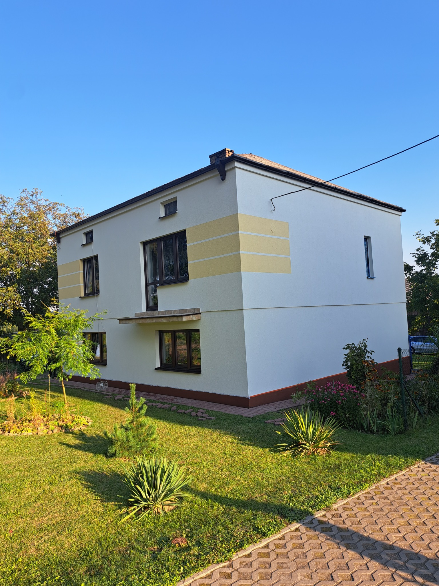 Dwukondygnacyjny dom z płaskim dachem, świeżo pomalowany na biało z poziomymi żółtymi pasami, widok z ogrodu w słoneczny dzień.