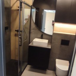 Wojciech Dutkiewicz - Nowoczesna łazienka z kabiną prysznicową typu walk-in, okrągłym lustrem i czarną armaturą na tle szarych płytek imitujących kamień, oświetlona punktowymi lampami sufitowymi.