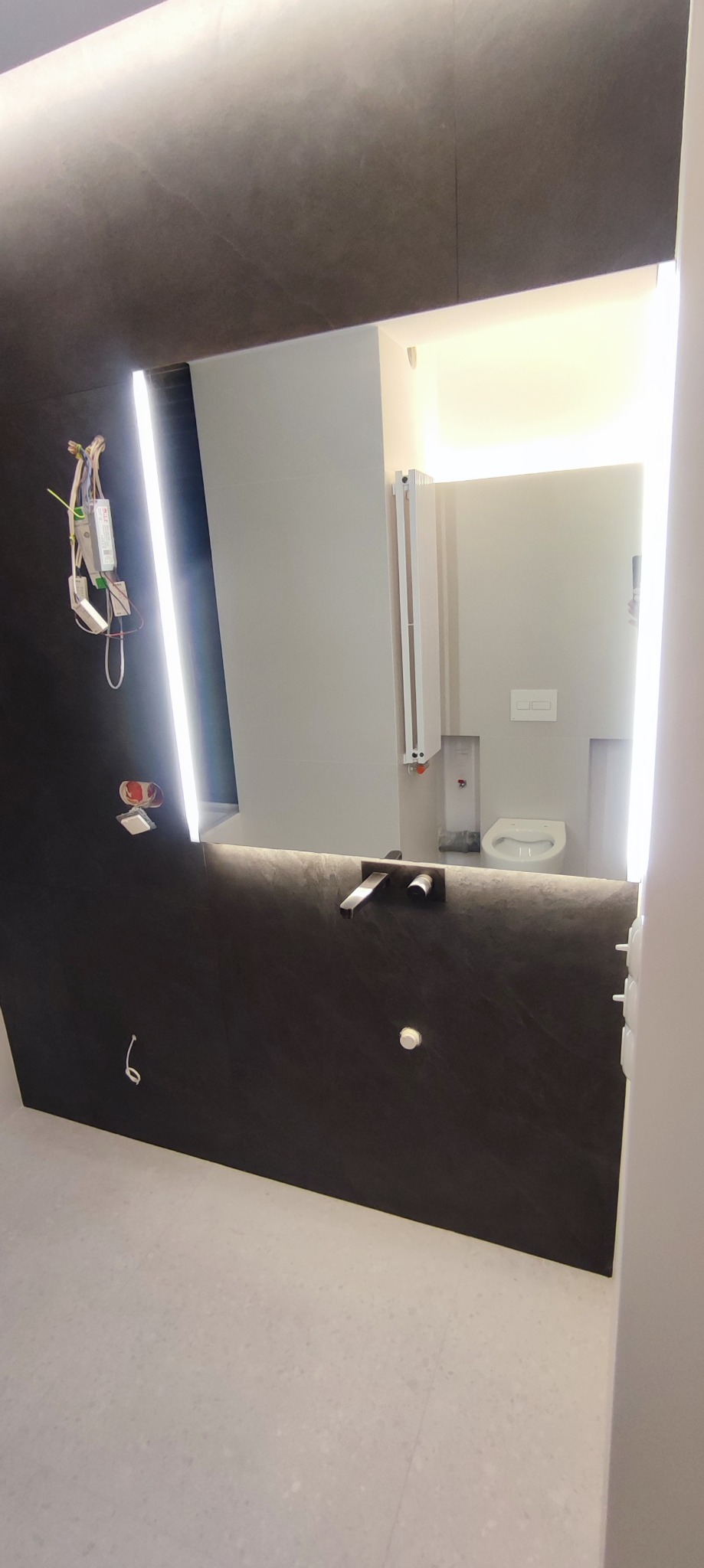 Ciemna ściana w łazience z podświetlanym lustrem, widoczne okablowanie i armatura, w odbiciu toaleta i grzejnik.