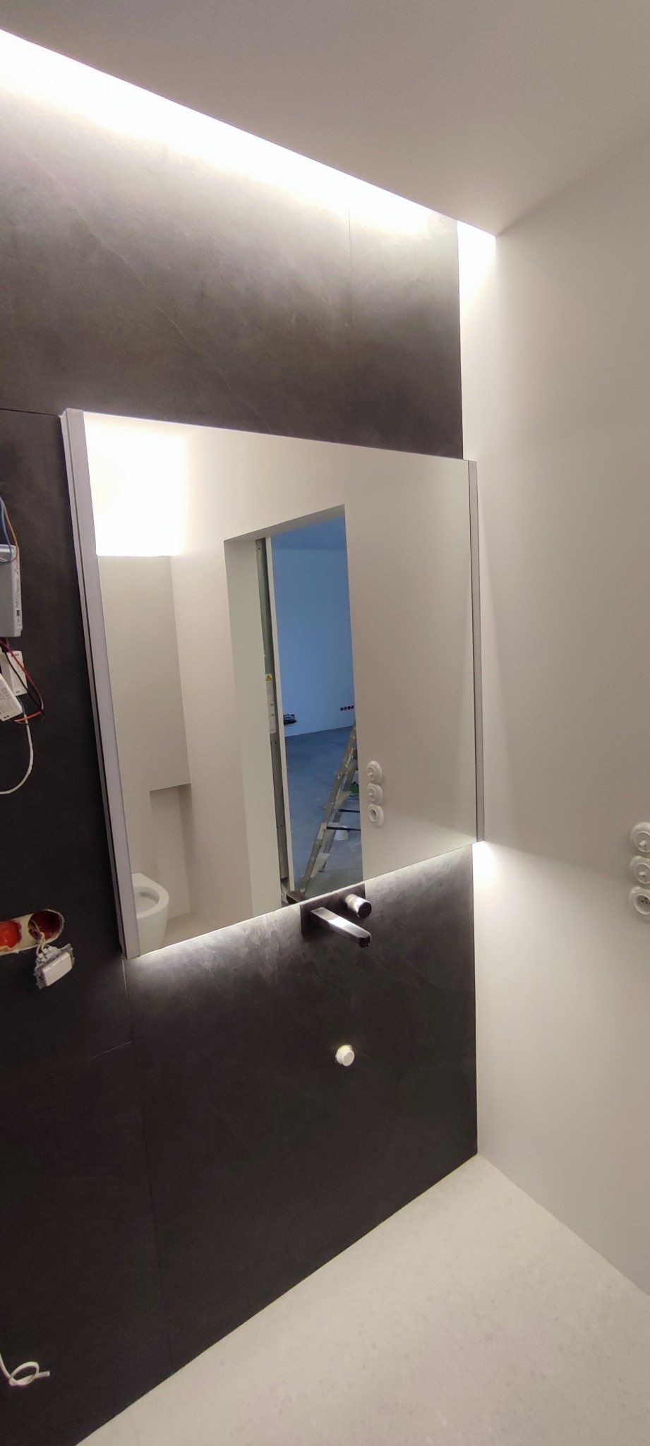 Nowoczesna łazienka z podświetlanym lustrem, ciemnoszara ściana z umywalką i baterią, widok na toaletę i drabinę w odbiciu lustra, elementy elektryczne widoczne po lewej stronie.