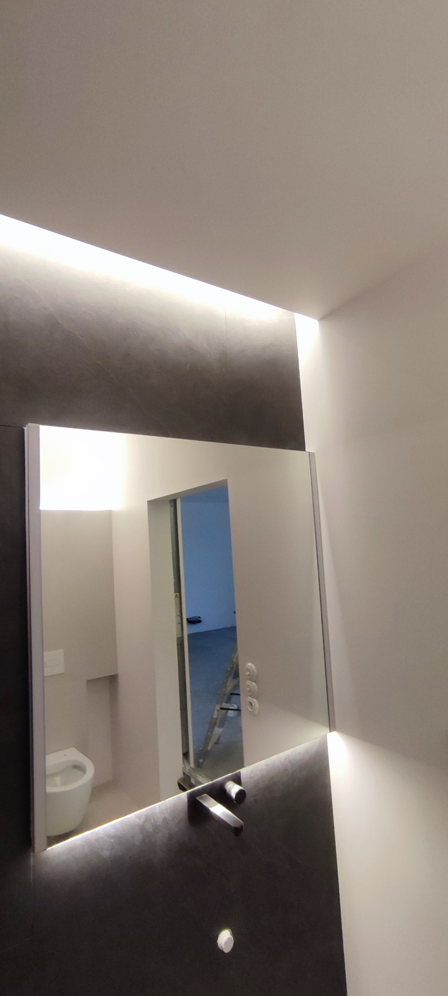 Nowoczesna łazienka z podświetlanym lustrem, ciemnoszarą ścianą z umywalką i widocznym w odbiciu fragmentem toalety i pomieszczenia w trakcie remontu z drabiną.