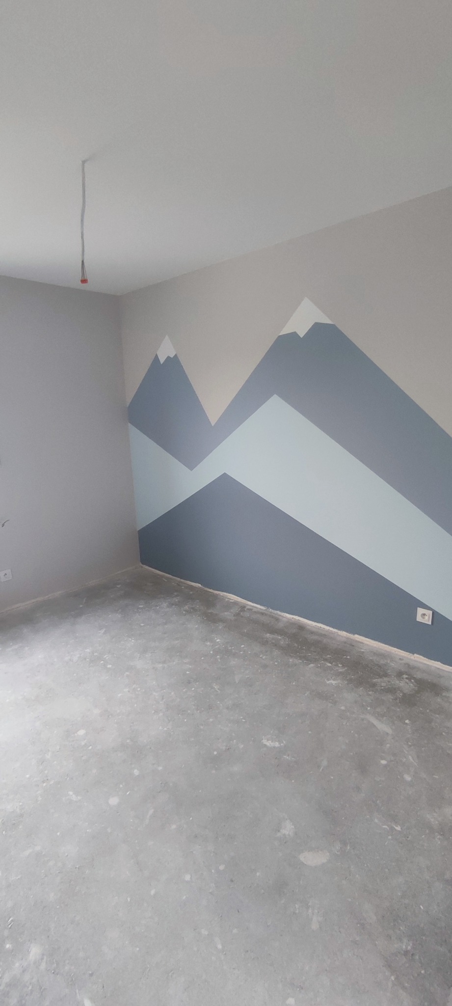 Pokój z szarą ścianą ozdobioną malunkiem gór w odcieniach szarości i beżu, surowy betonowy podkład pod podłogę, wystające przewody elektryczne z sufitu.