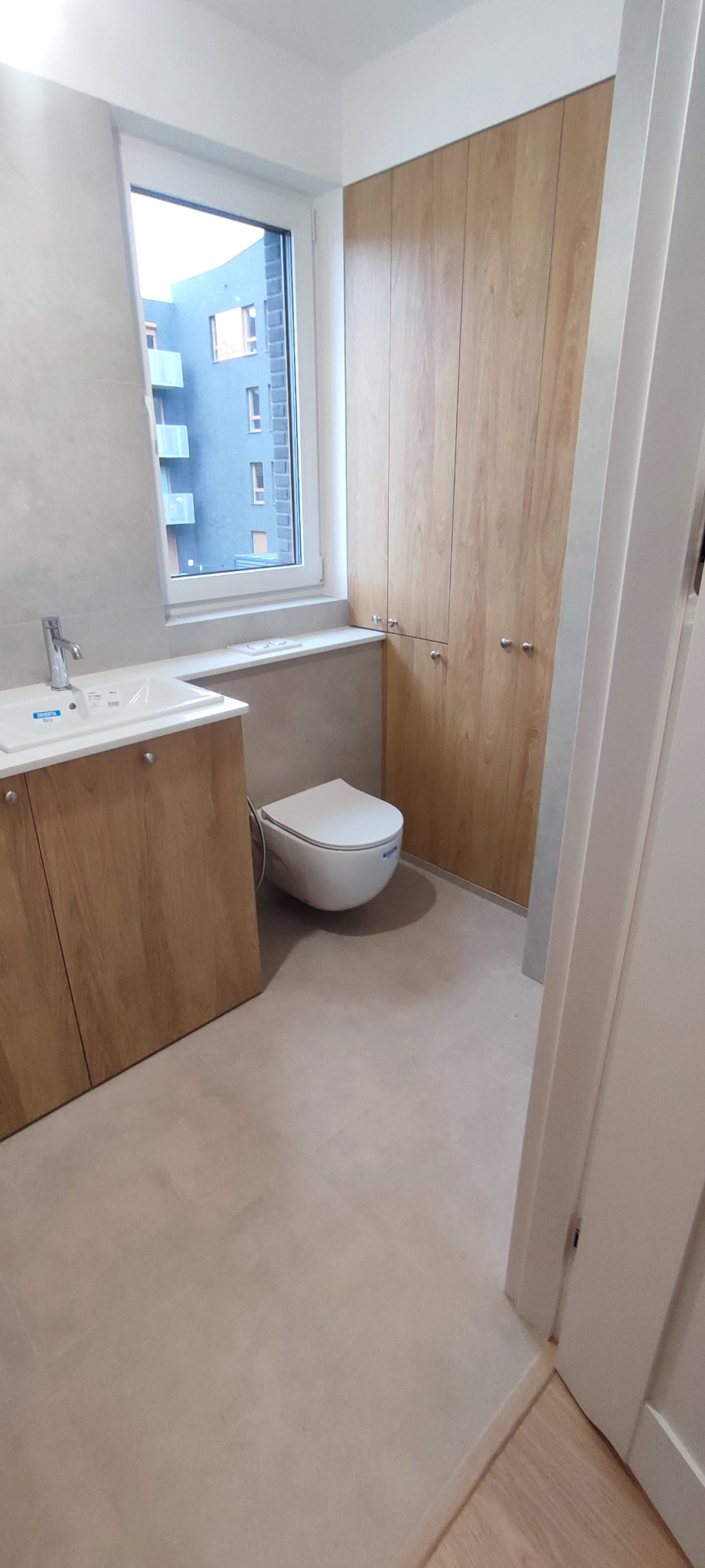 Nowoczesna łazienka z podwieszaną toaletą, szafką pod umywalką z drewnopodobnym frontem i wysoką szafą wnękową z jasnego drewna, widok na budynek z okna.