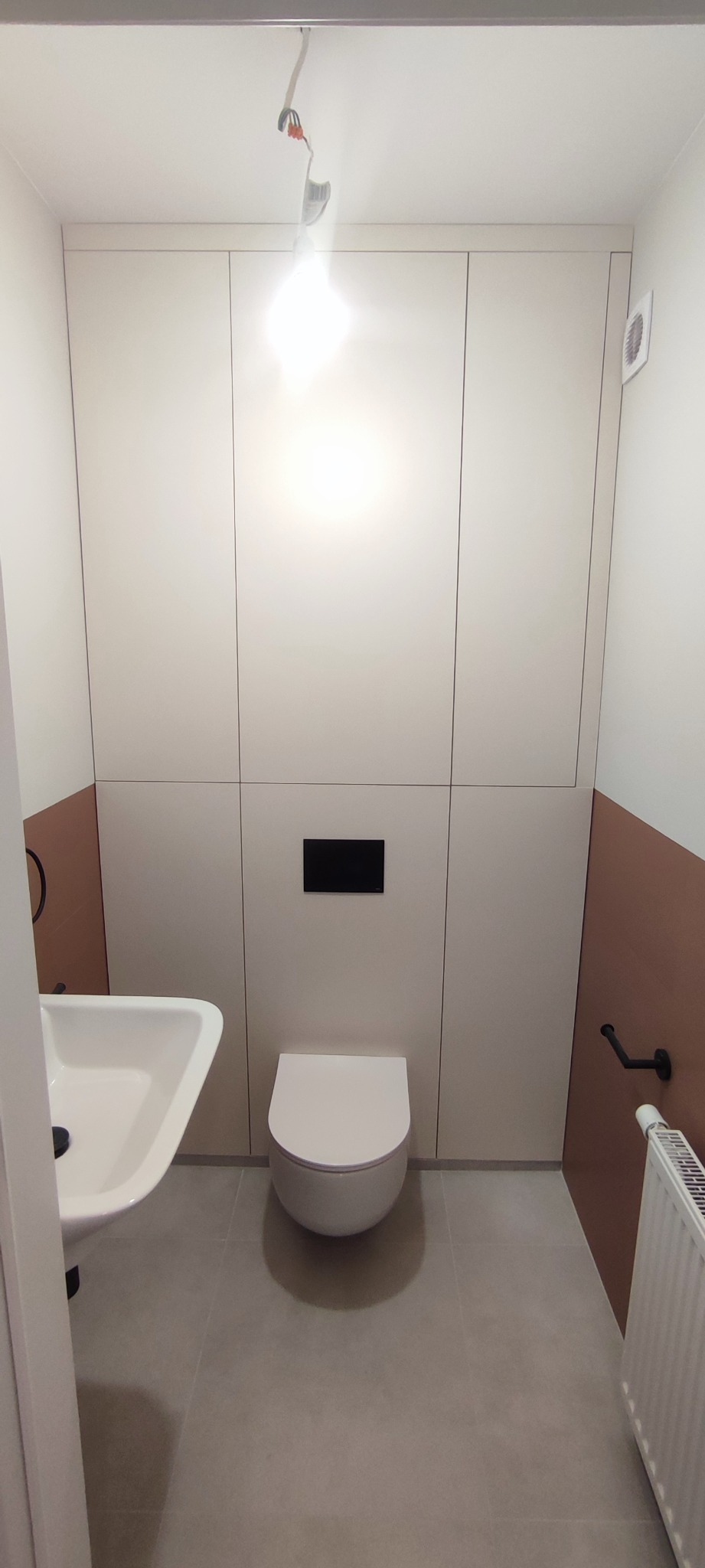 Nowoczesna łazienka z wiszącą toaletą, umywalką i zabudową meblową w kolorze kremowym, akcentowana brązowym pasem na ścianie i czarnymi dodatkami, oświetlona surową żarówką wiszącą.